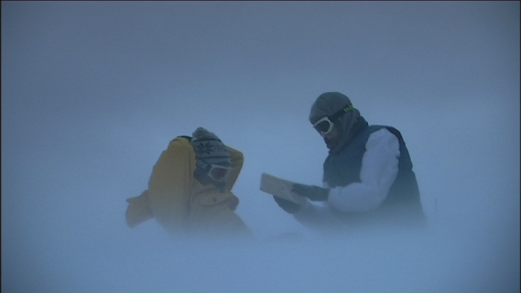 Two men wearing hoods and masks, one reading a book