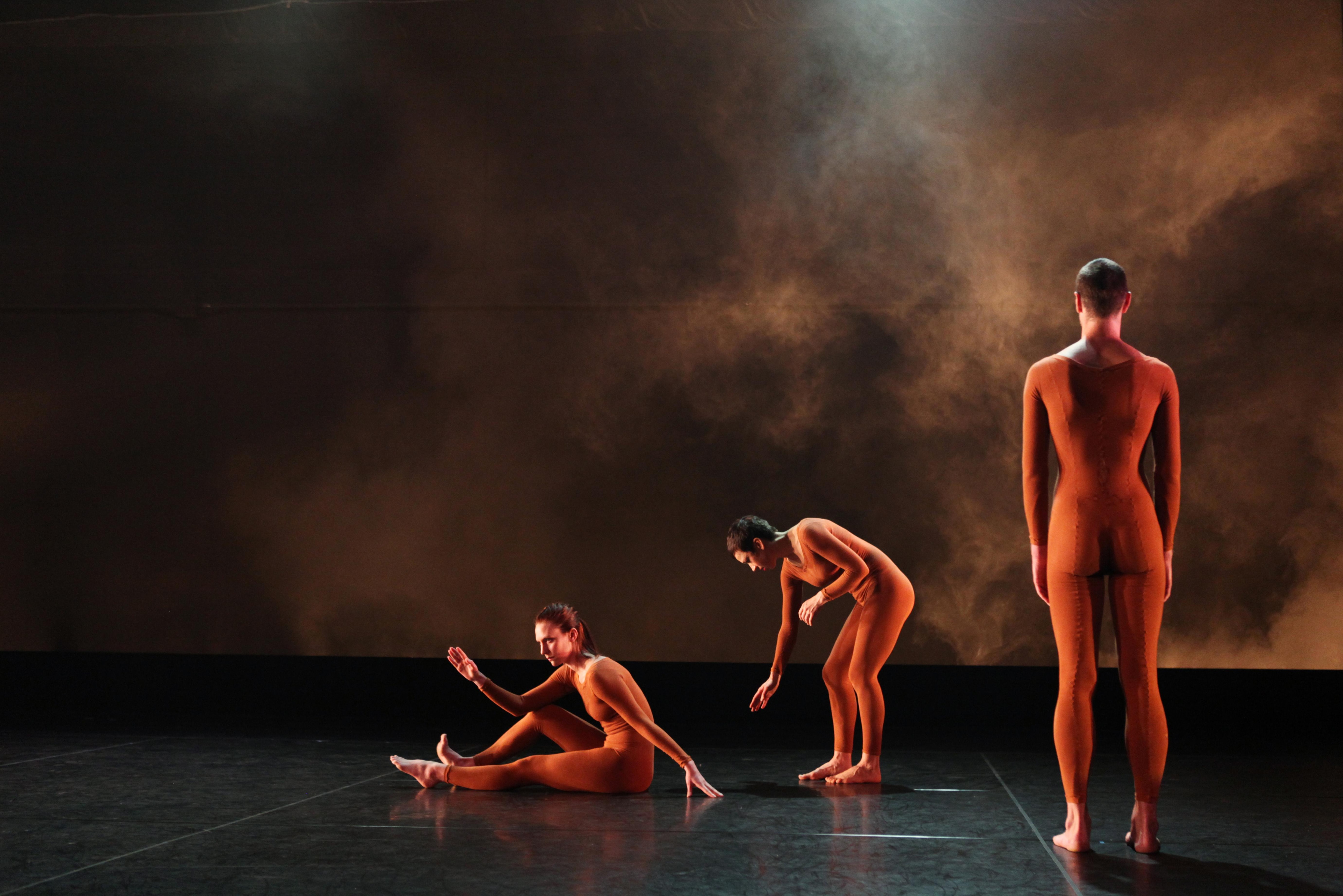 Three dancers on stage, the first one is sitting with his legs on the floor and one arm on his knee, the second one is leaning down, and the third one is standing from behind