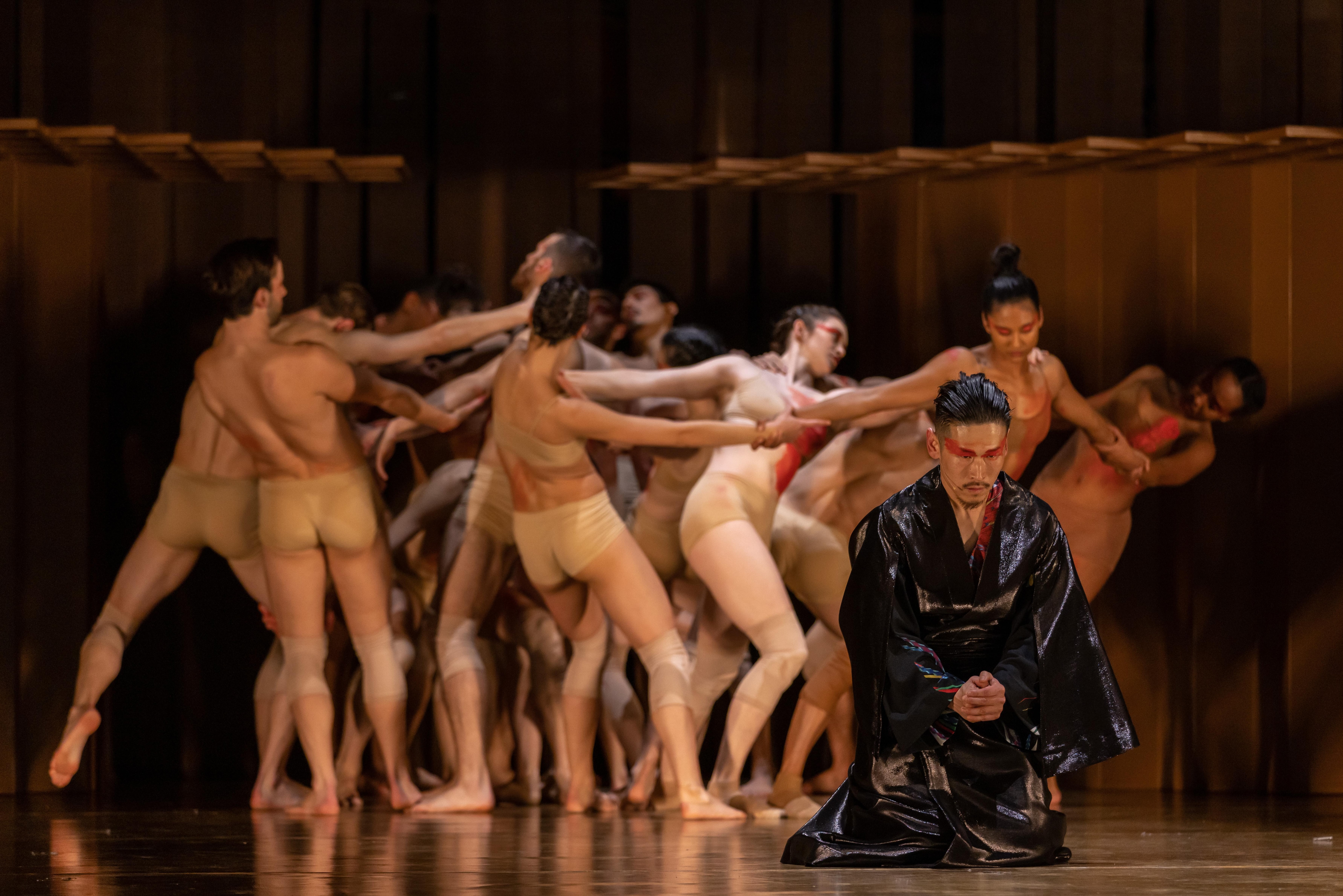 Danseur avec du maquillage rouge sur les yeux, en position de prière. En arrière-plan, des danseurs vêtus de sous-vêtements beiges sont regroupés et se touchent. 