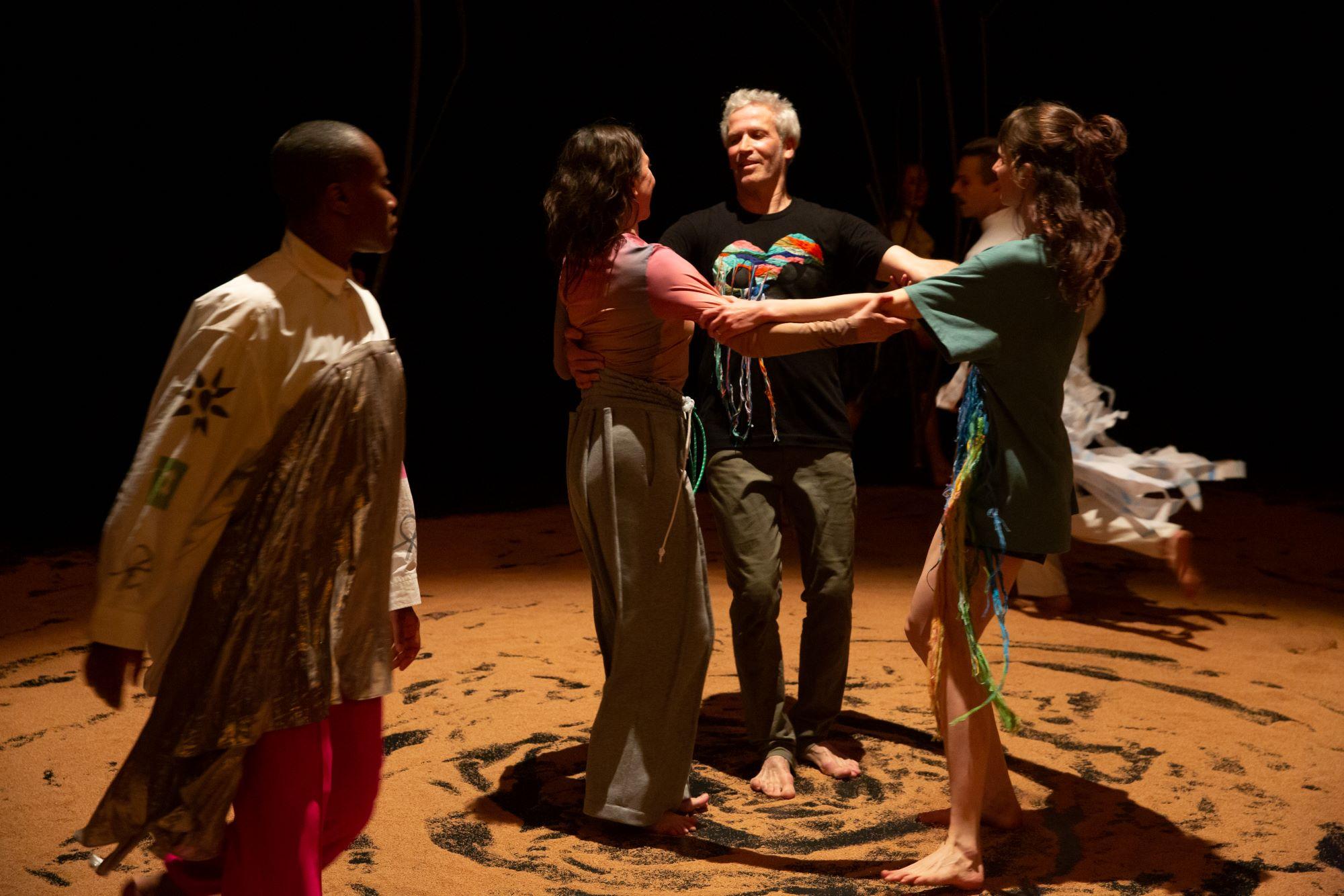 Trio of dancers holding each other's arms in a circle, feet in the sand