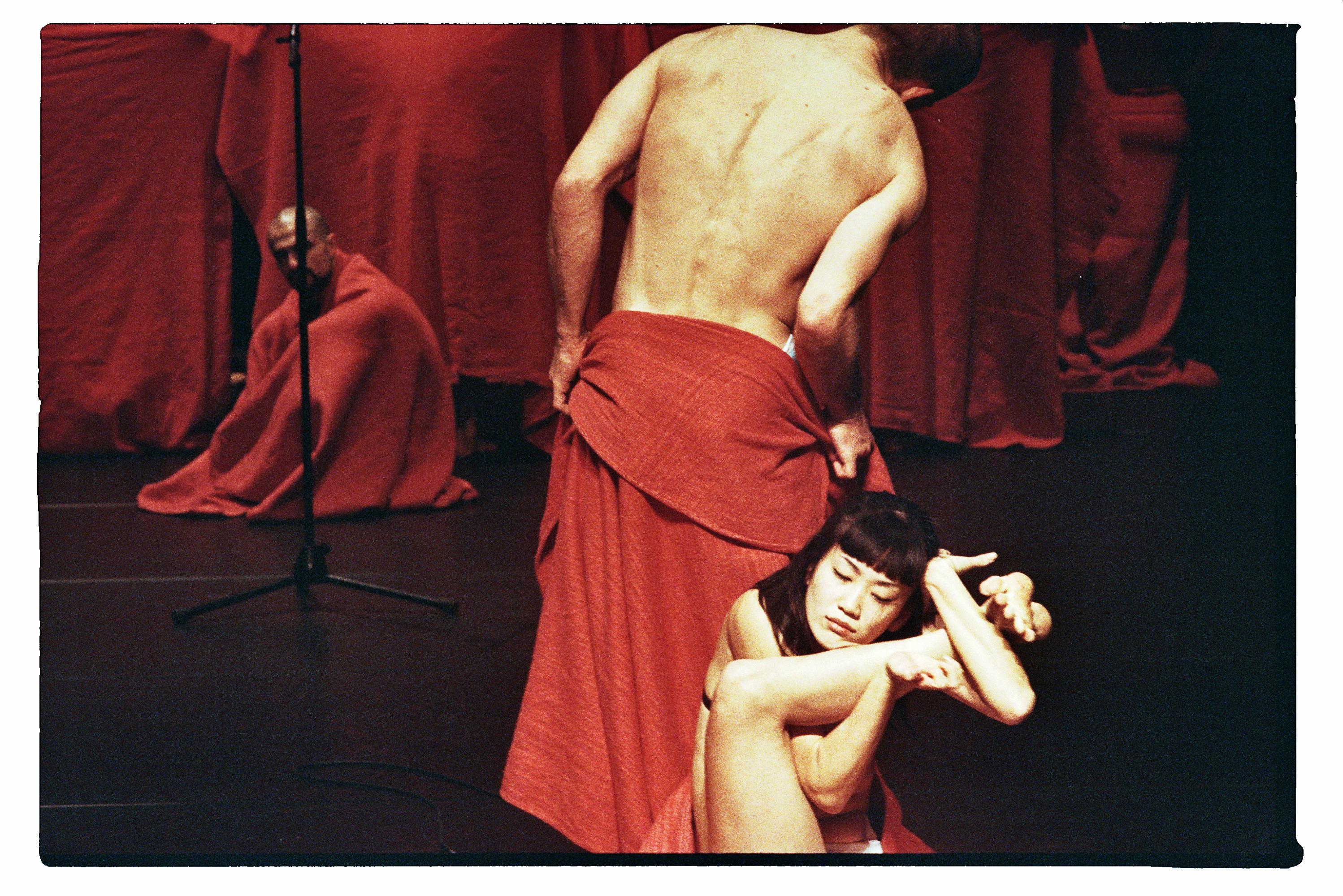 A dancer with a red blanket around his waist and another dancer sitting acrobatically in front of him