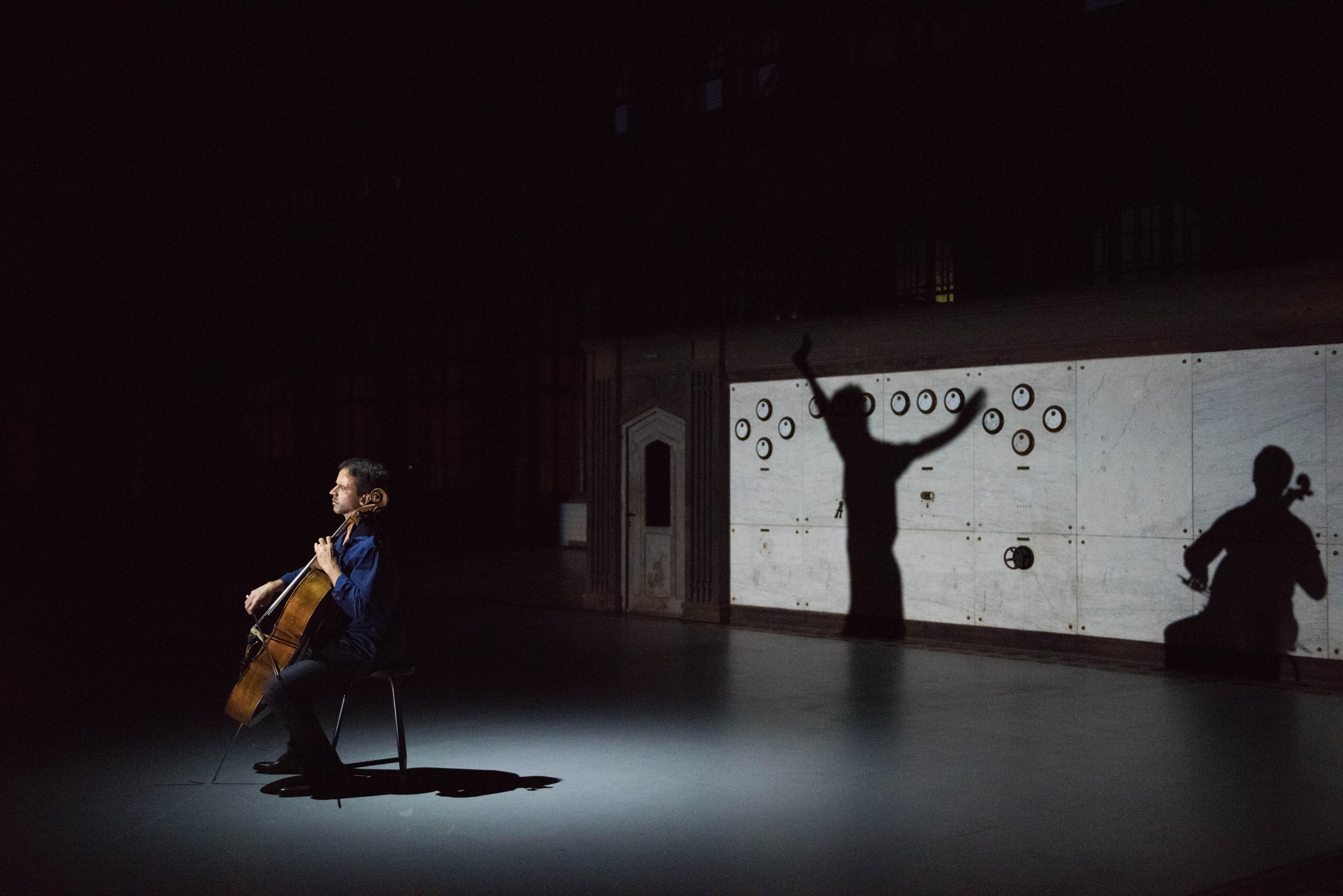 Cellist Jean-Guihen Queyras in front of a concrete wall, with the shadow of two dancers.
