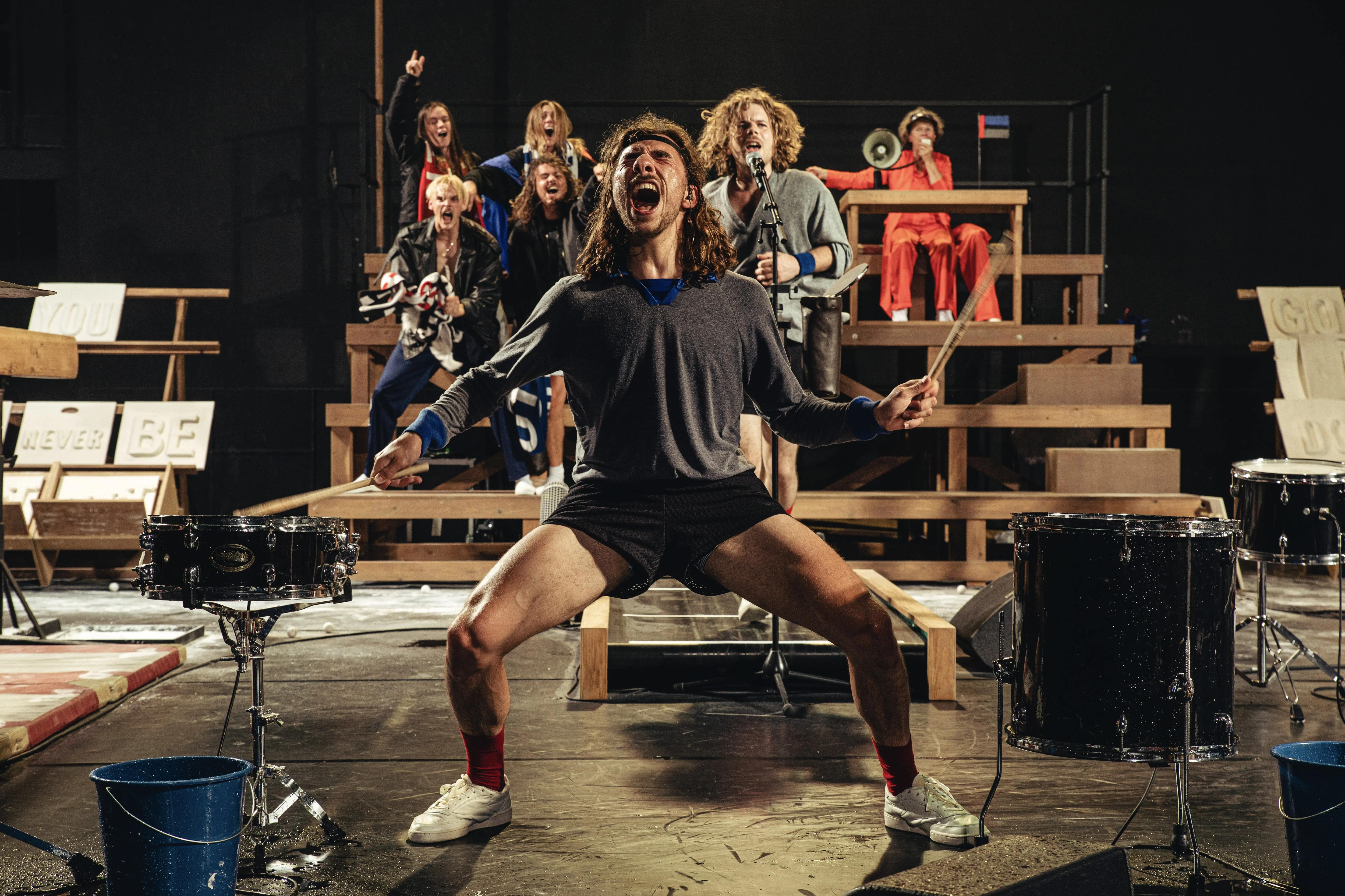 Six people shouting on the bleachers. A man shouting in the front of of the stage, legs spread apart 