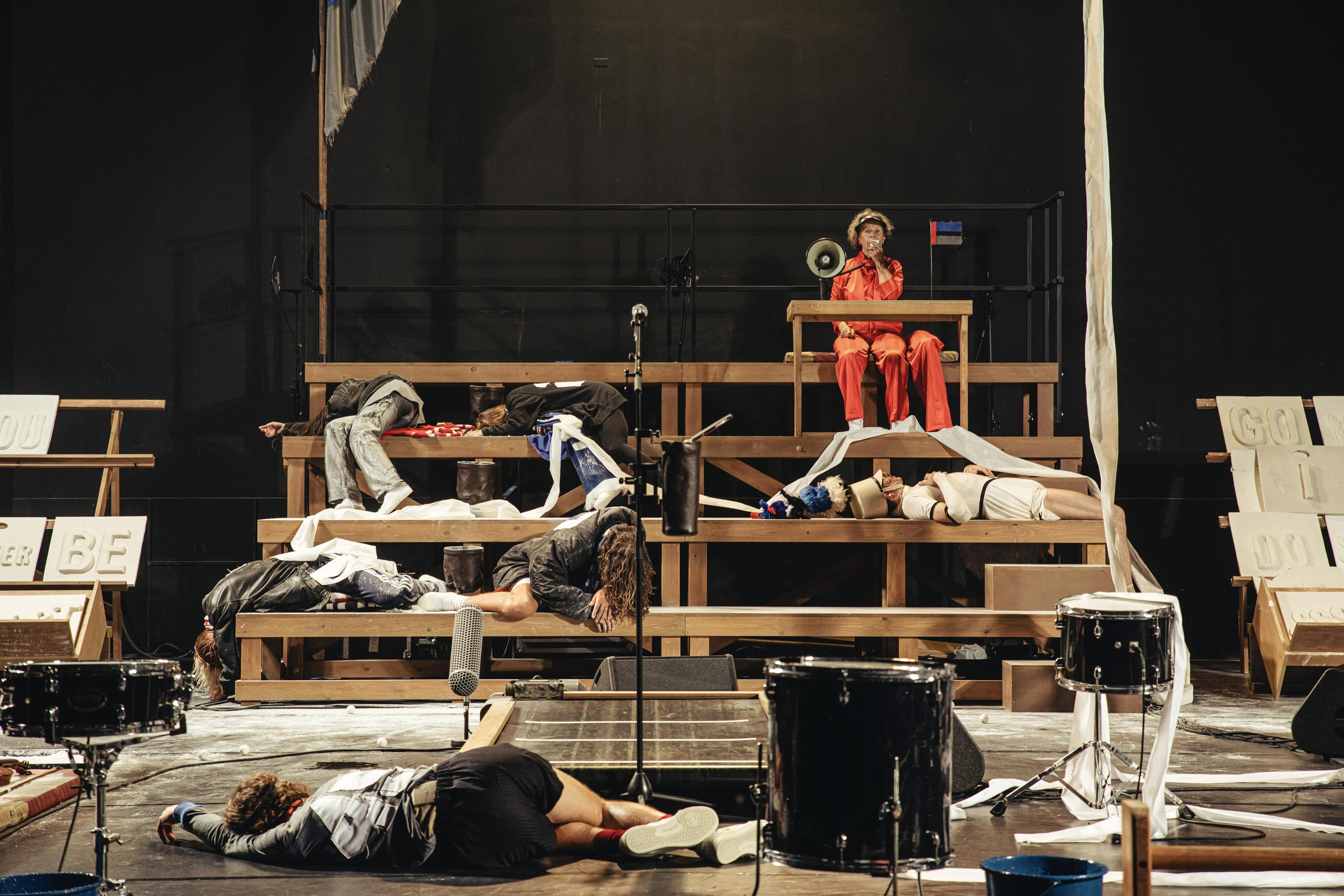 Six people shouting on the bleachers. A man shouting in the front of of the stage, legs spread apart 