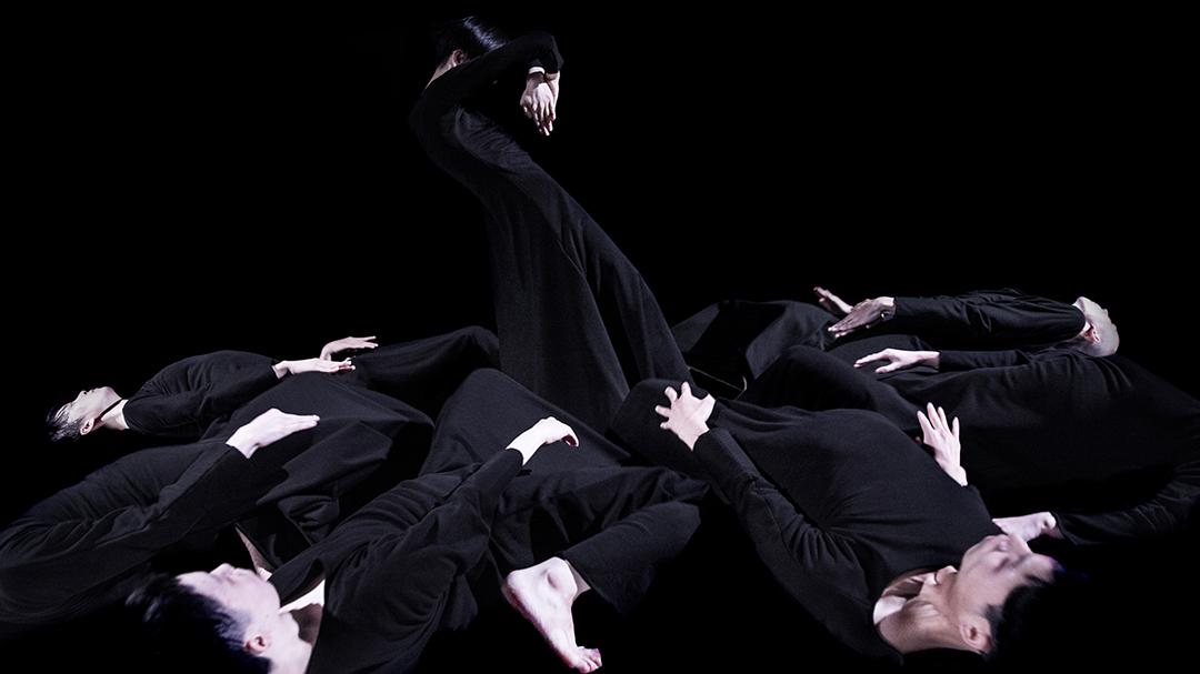 Danseur émergeant d’un groupe de danseurs en cercle, tous vêtus de noir.