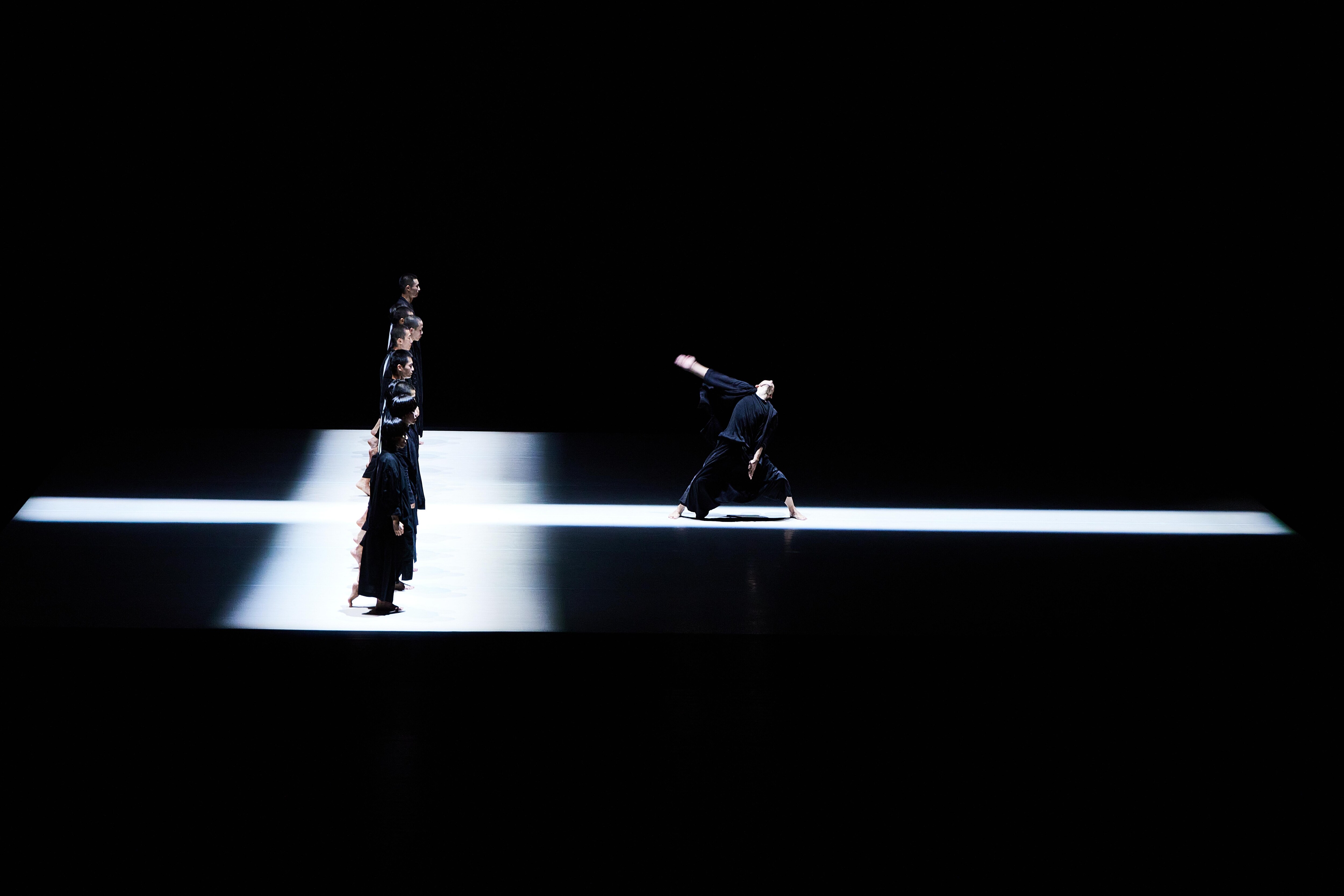 danseurs alignés sur un passage de lumière blanche, faisant face à un autre danseur, seul sur une ligne de lumière perpendiculaire.