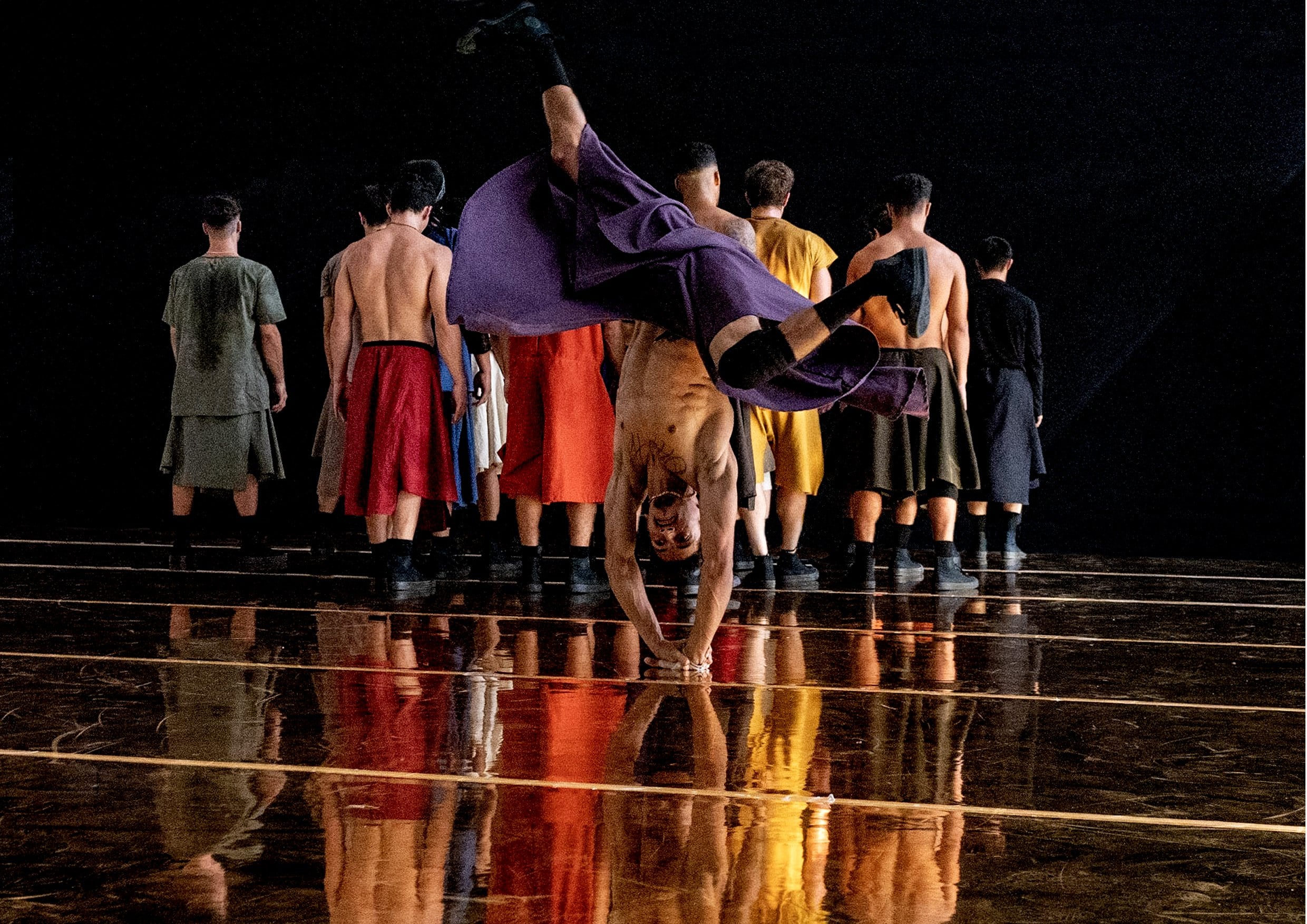 Danseur en paréo violet faisant une acrobatie sur le mains, groupe de danseurs de dos au fond.