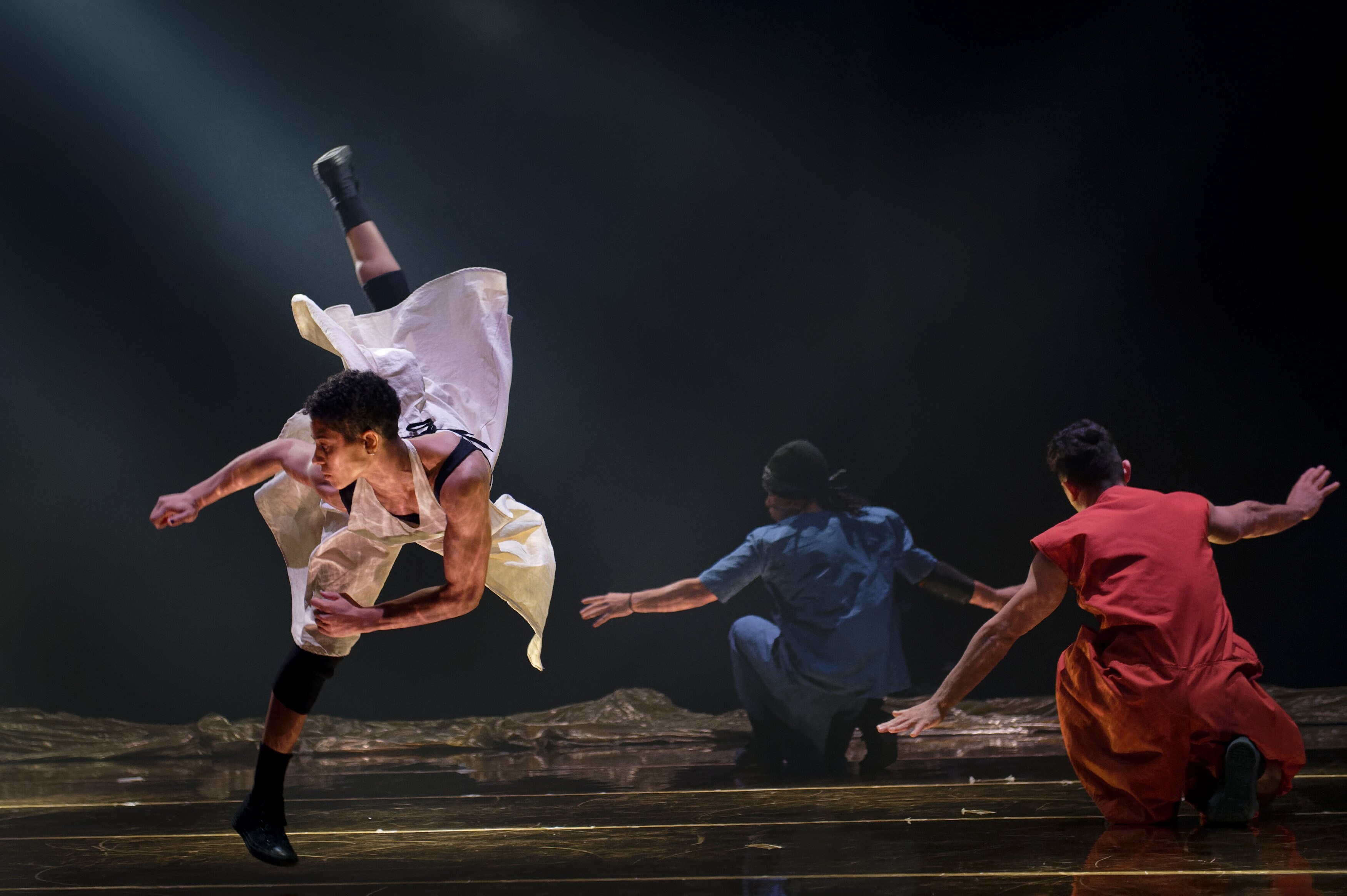 Une danseuse dans une robe blanche effectuant un saut tourné, un danseur en bleu et un en rouge par terre en arrière-plan 