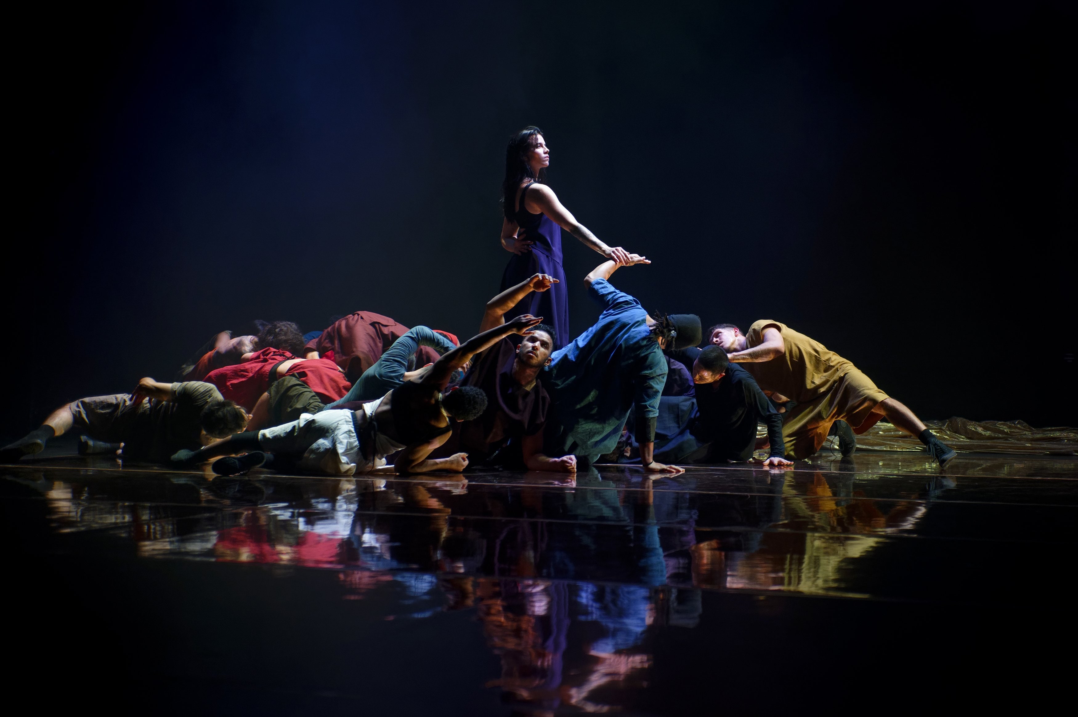 Dancers in a group on the floor in colorful clothes, a single dancer standing in the middle.