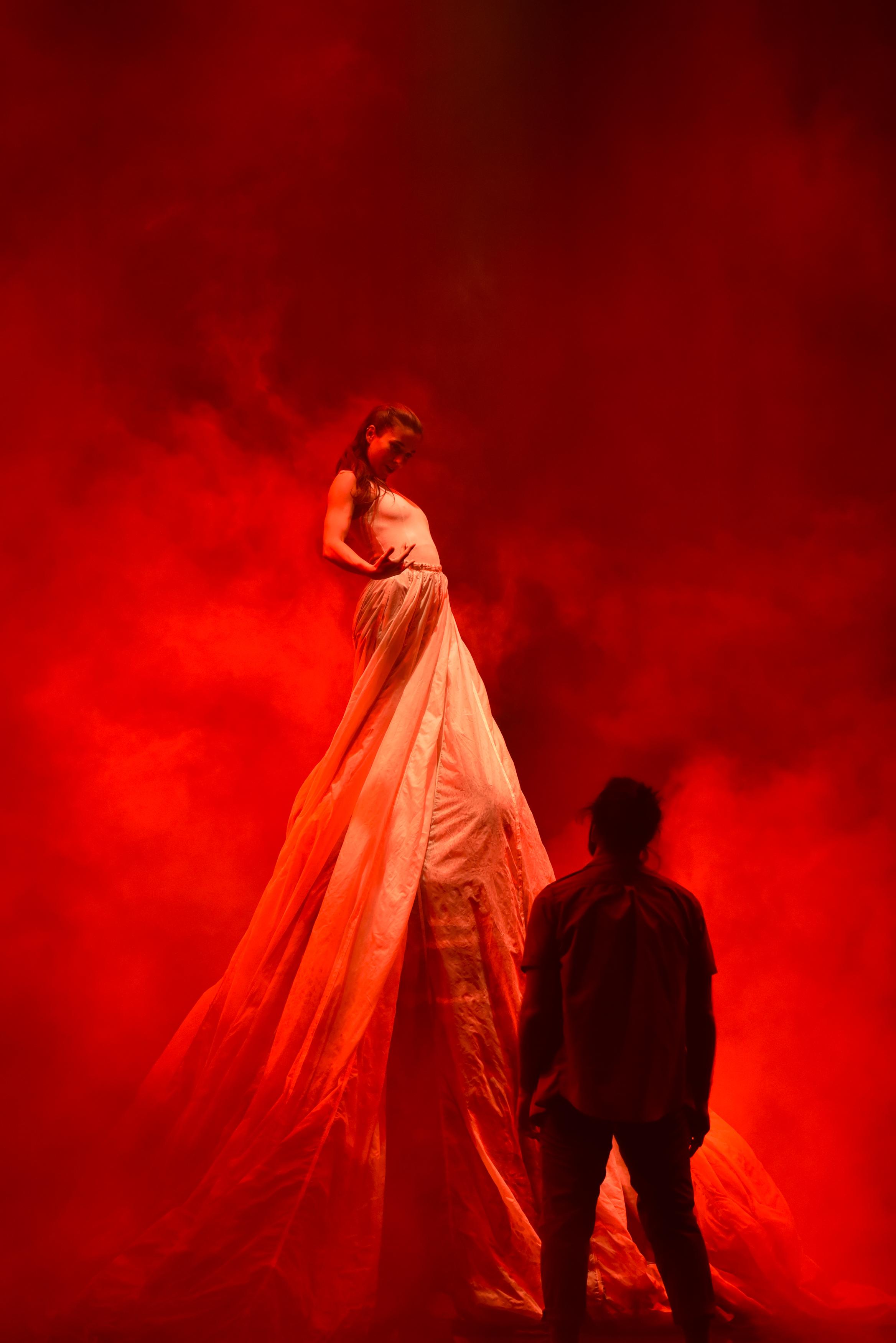 Homme de dos regardant une femme dans une grande robe faisant deux fois sa hauteur, ambiance rougeâtre. 