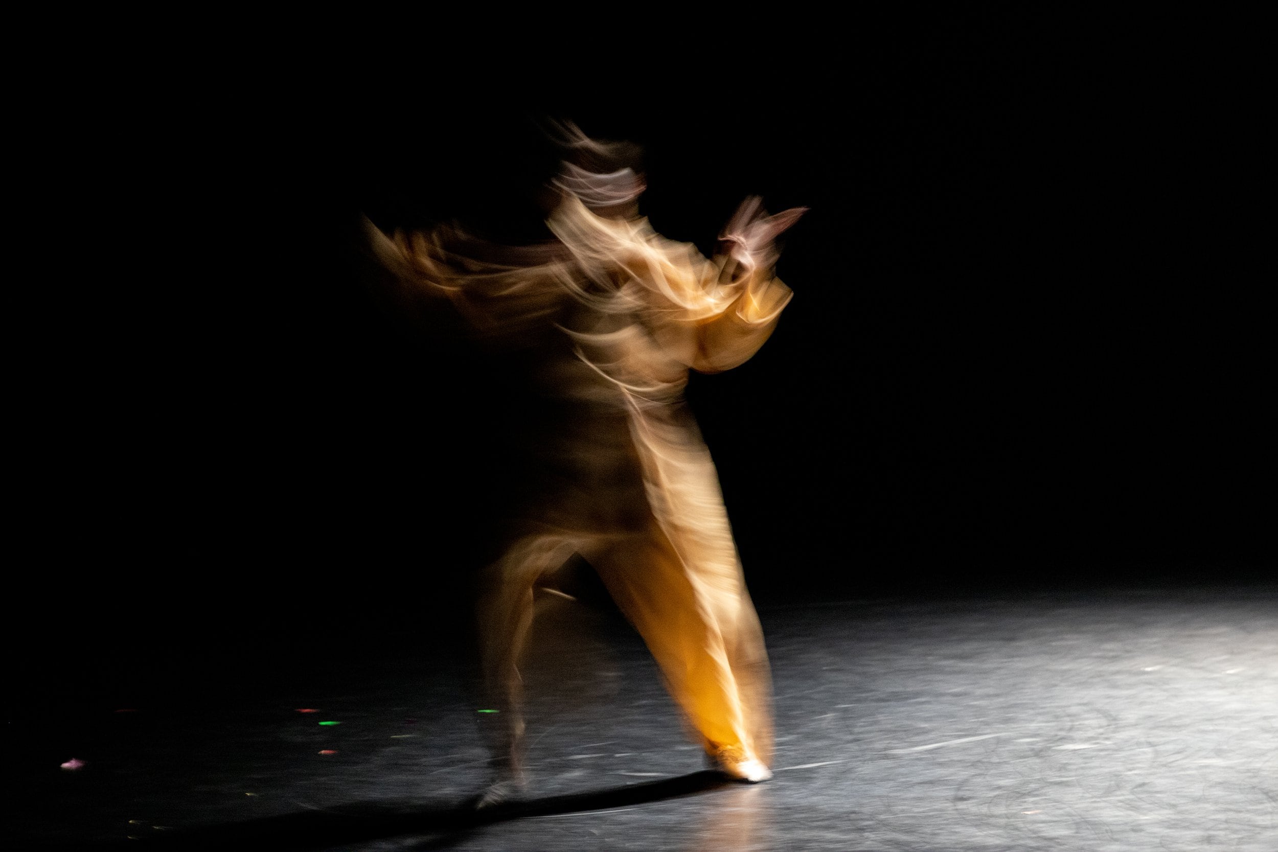 A blurry motion image capture of a pale orange figure in a black theater. The arms are outstretched. The image is reminiscent of flickering flames. 