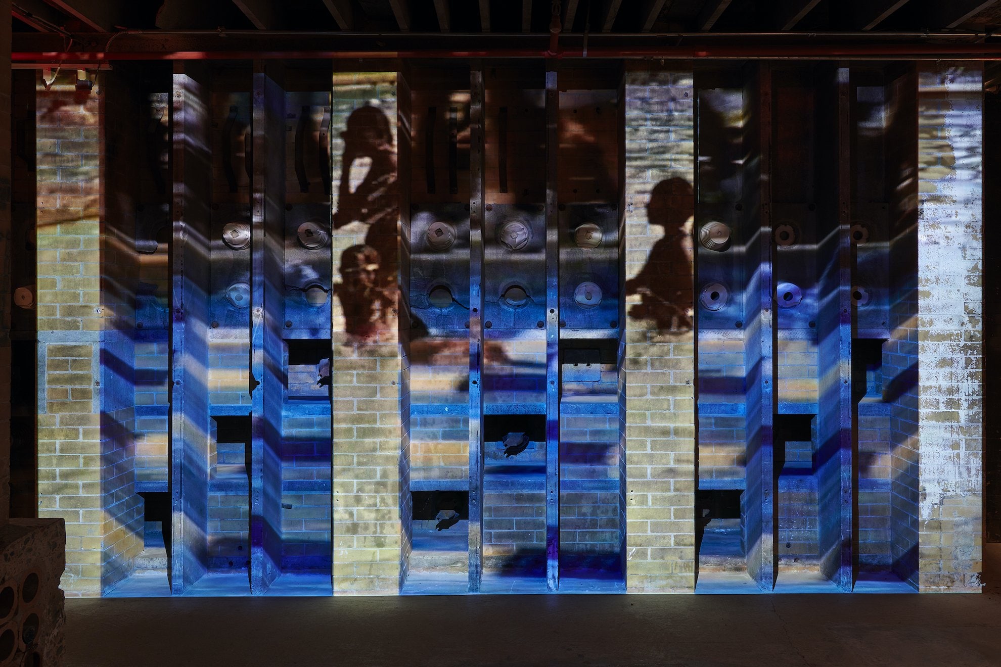 A video projection onto a series of recessed brick walls illuminates a Black woman and children in a swimming pool amidst blue waves.  