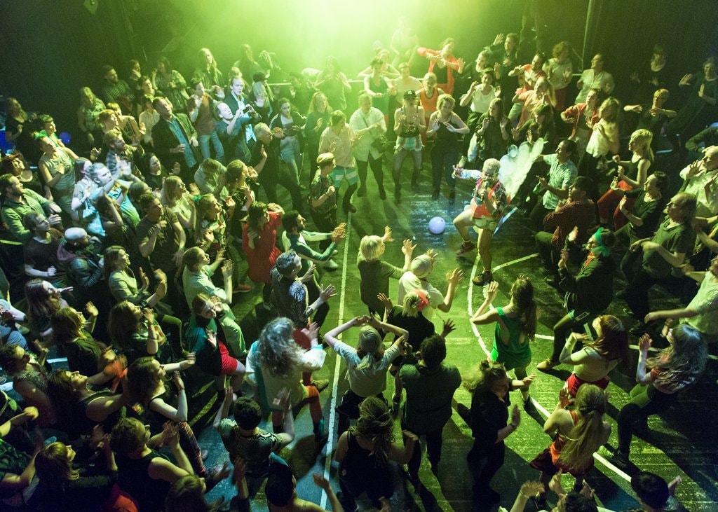 A large group of people in a gymnasium surround the artist, dressed in a costume evoking bubbles and high-energy aerobics, who guides them in movement. The participants are bathed in a green-glowing light and photographed from above.  