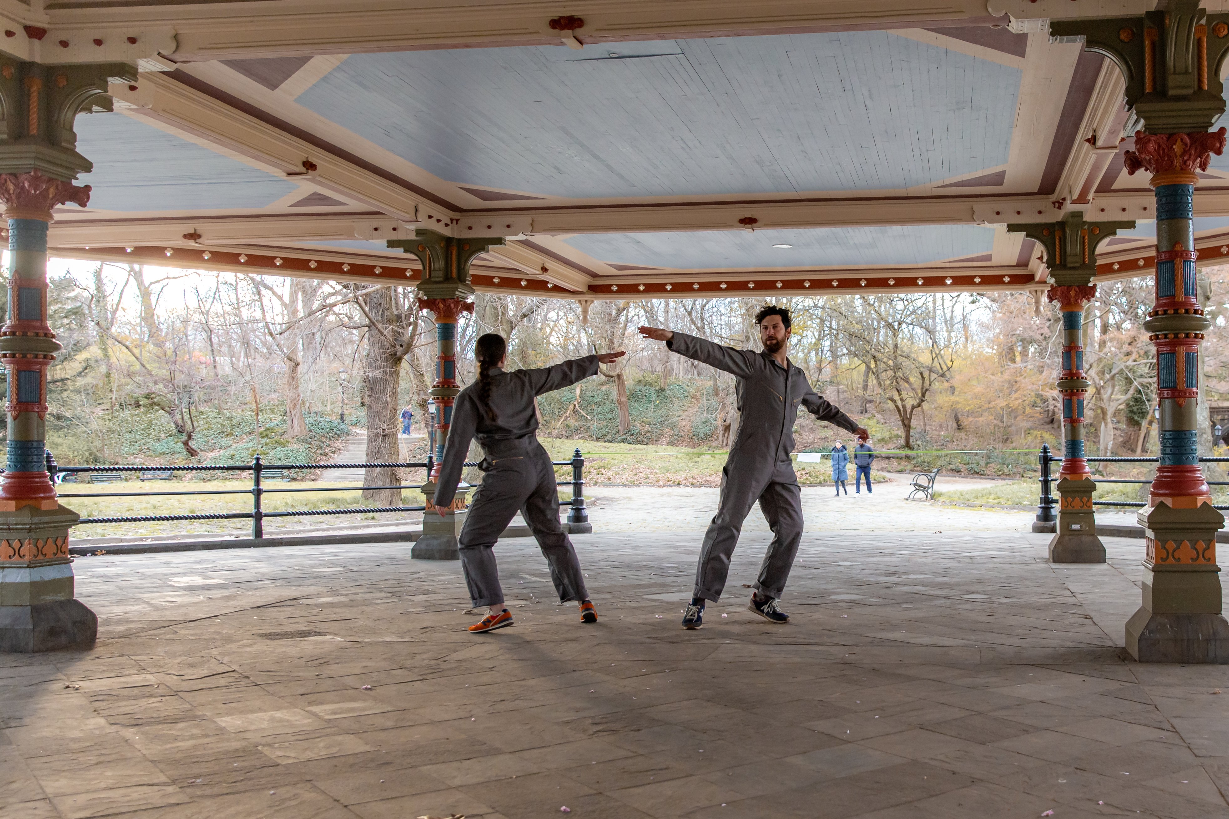 Under an ornate pavilion with trees off in the distance, two dancers in gray jumpsuits are caught turning with an arm extended to their side.  