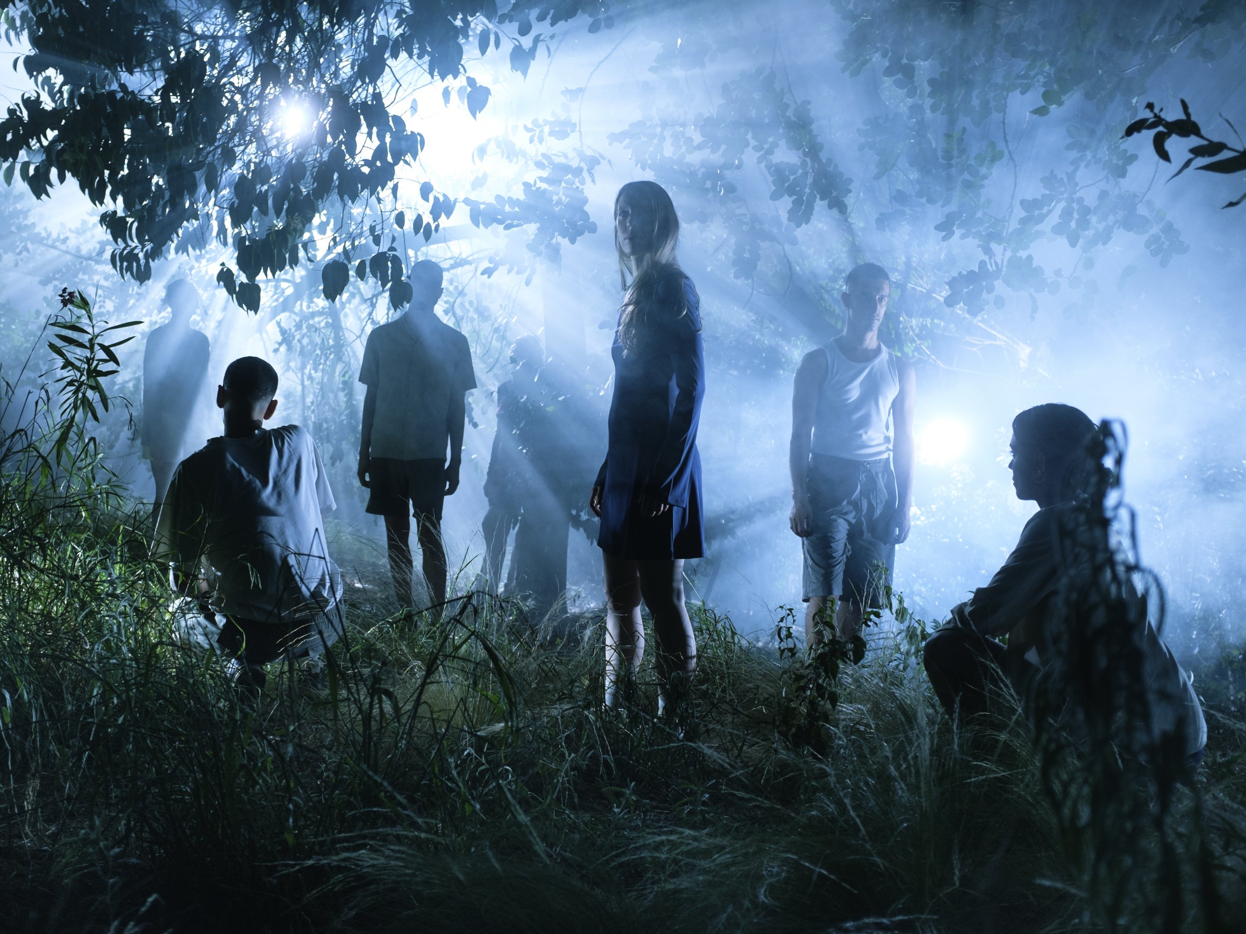 seven people, including two squatting and the others standing, in the dark forest.
