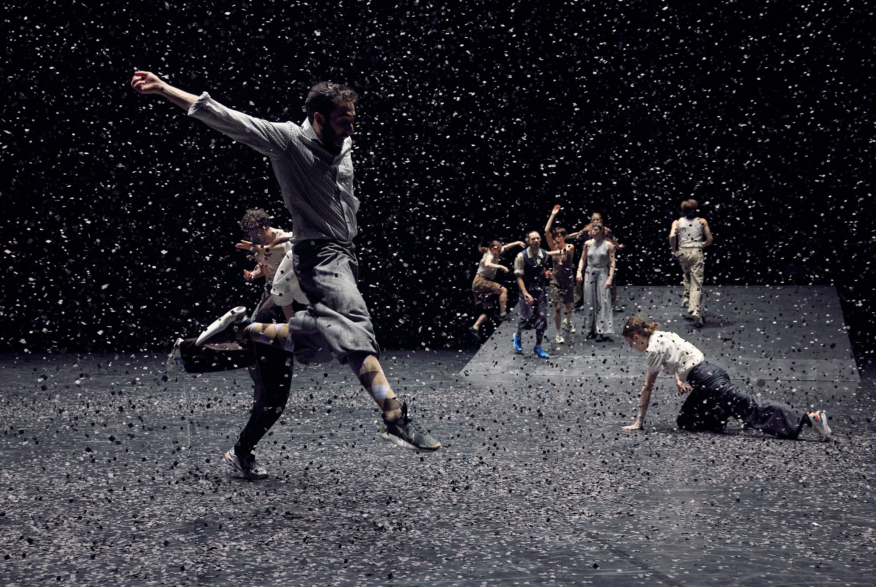 A group of dancers in the background, three dancers in the foreground, including one in the air under confetti