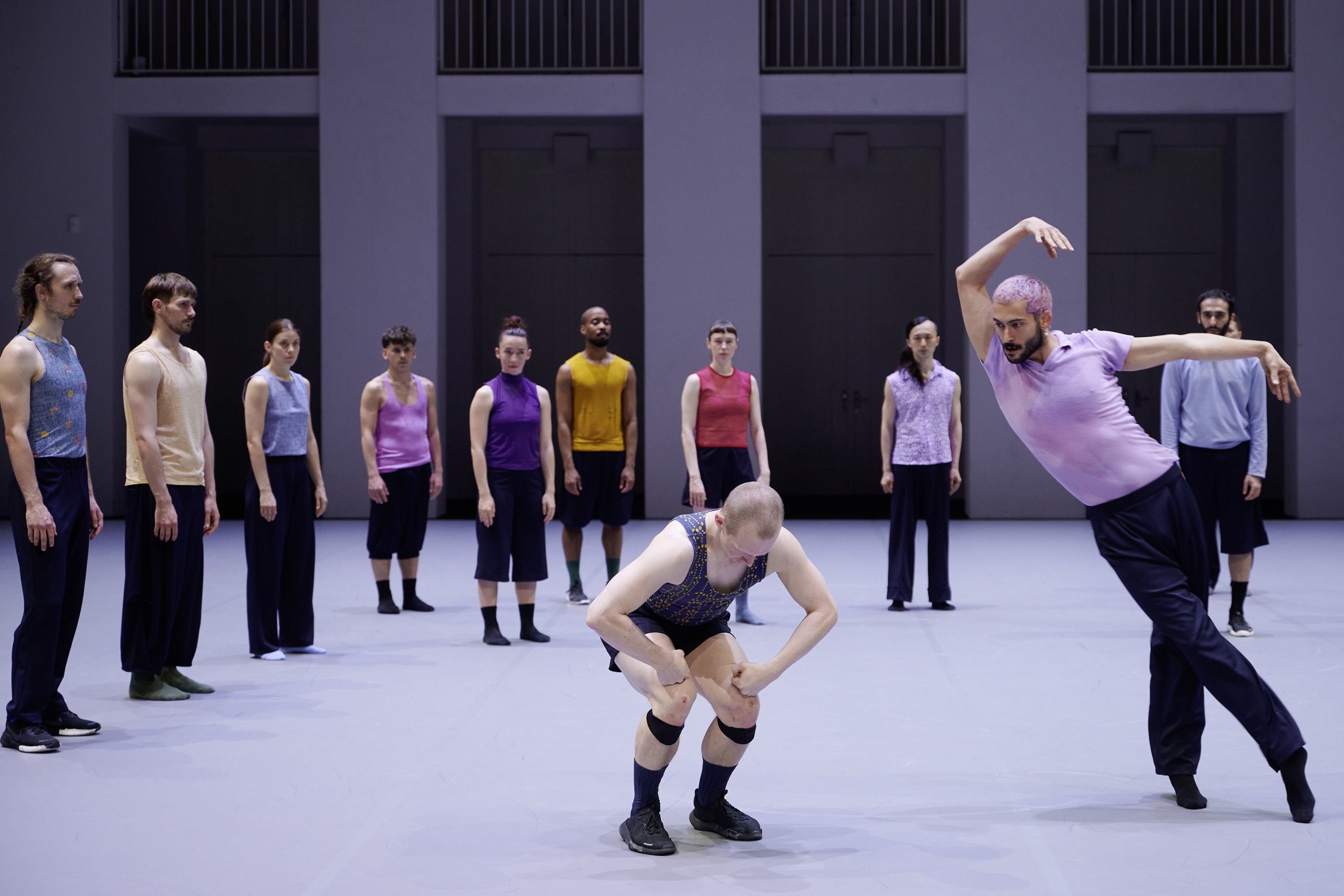Two dancers, one of whom is curled up, and the other dancers are watching them in the background