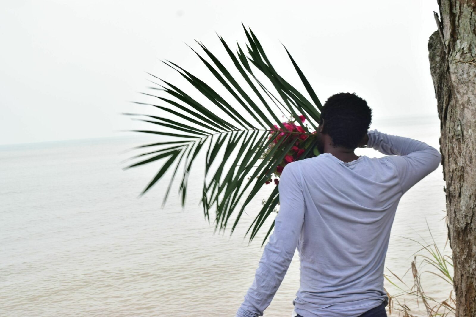 A man from behind looking at the sea with a palm leaf in his hands