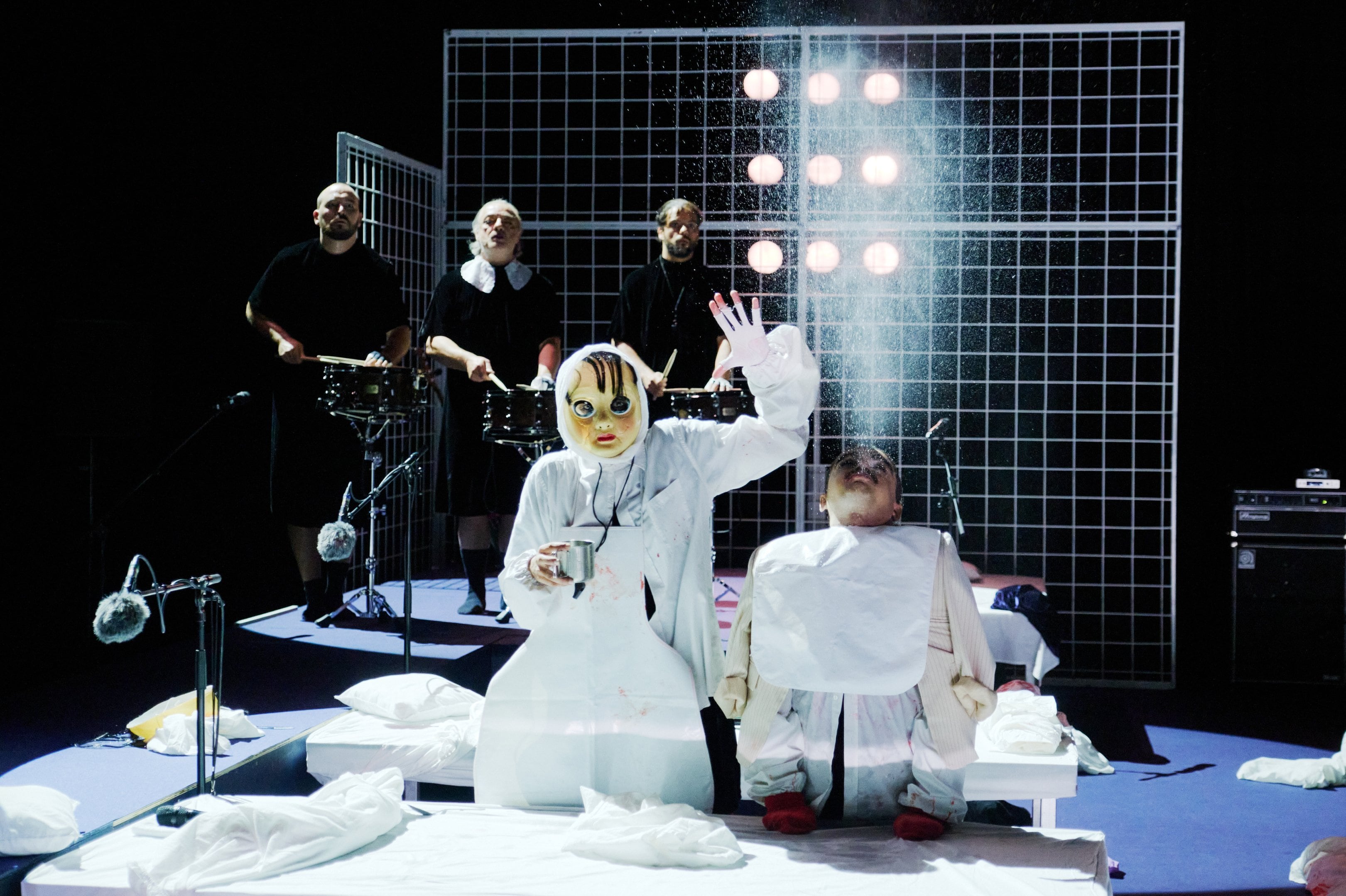 deux personnages en costumes blancs, l'un masqué et l'autre ressemblant à une marionnette, devant des musiciens