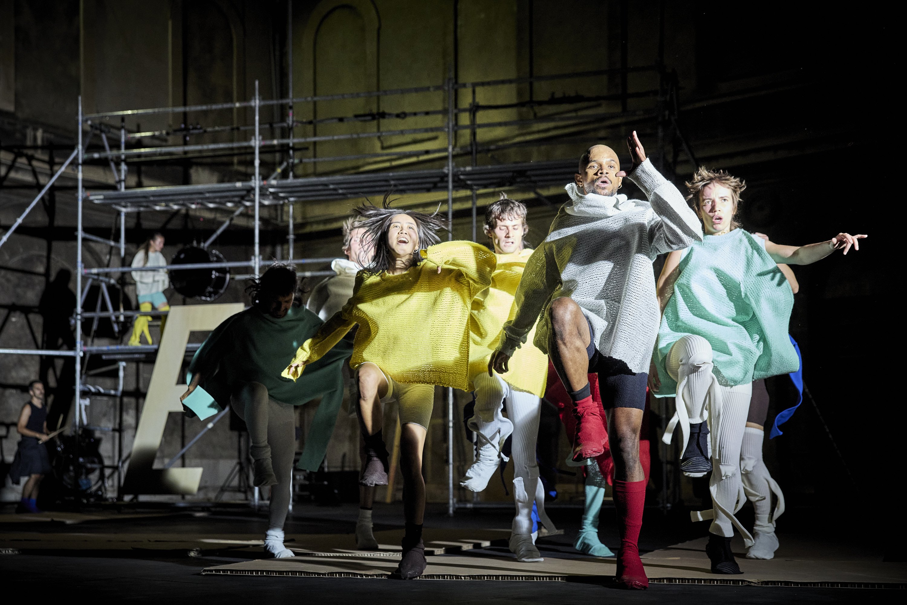 groupe de danseurs vêtus de hauts en tricot amples et colorés et de bas variés se produisent sur une scène, en mouvement avec des visages expressifs