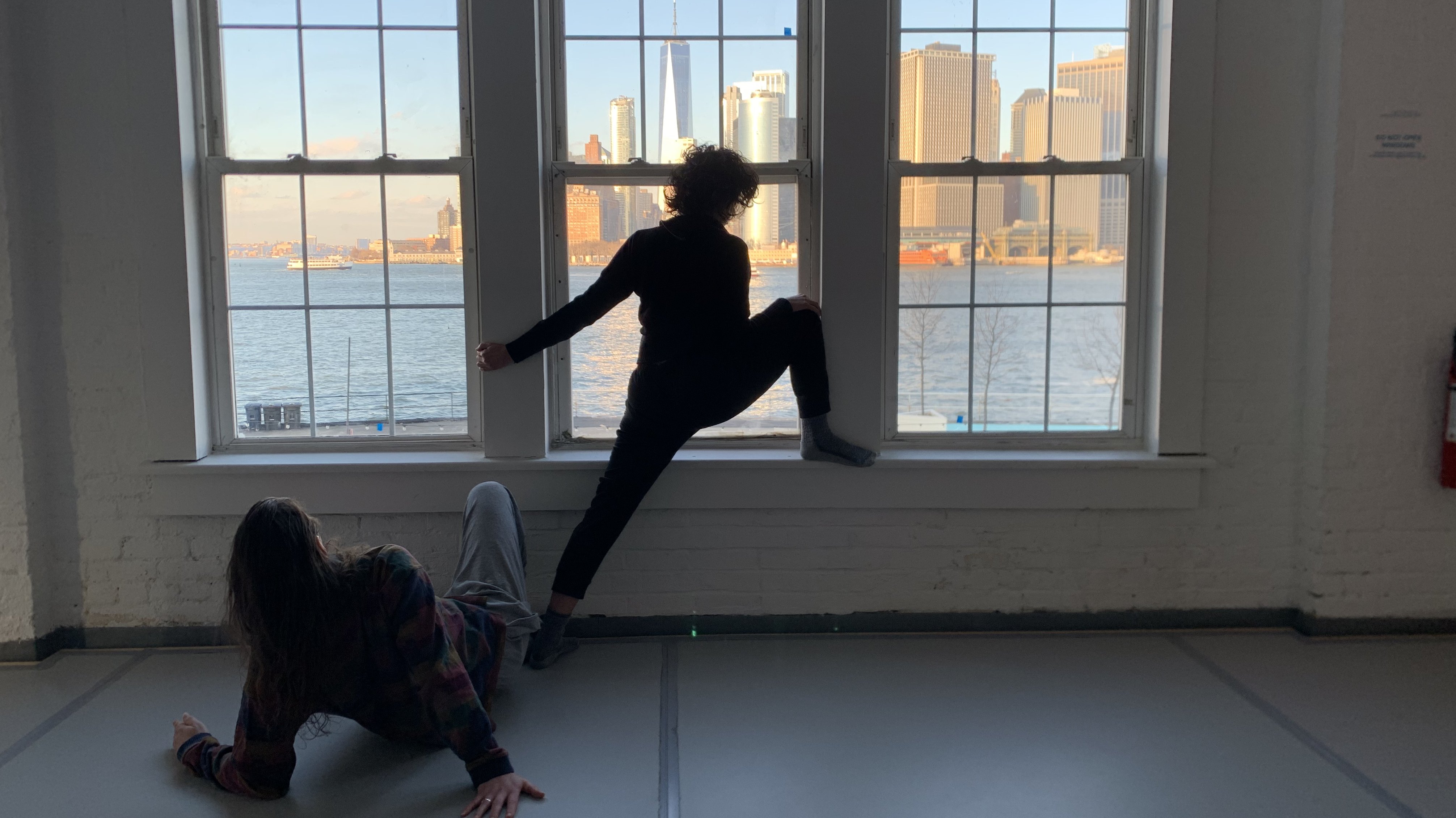 Two dancers stretching in front of a window with a view of  the New York landscape