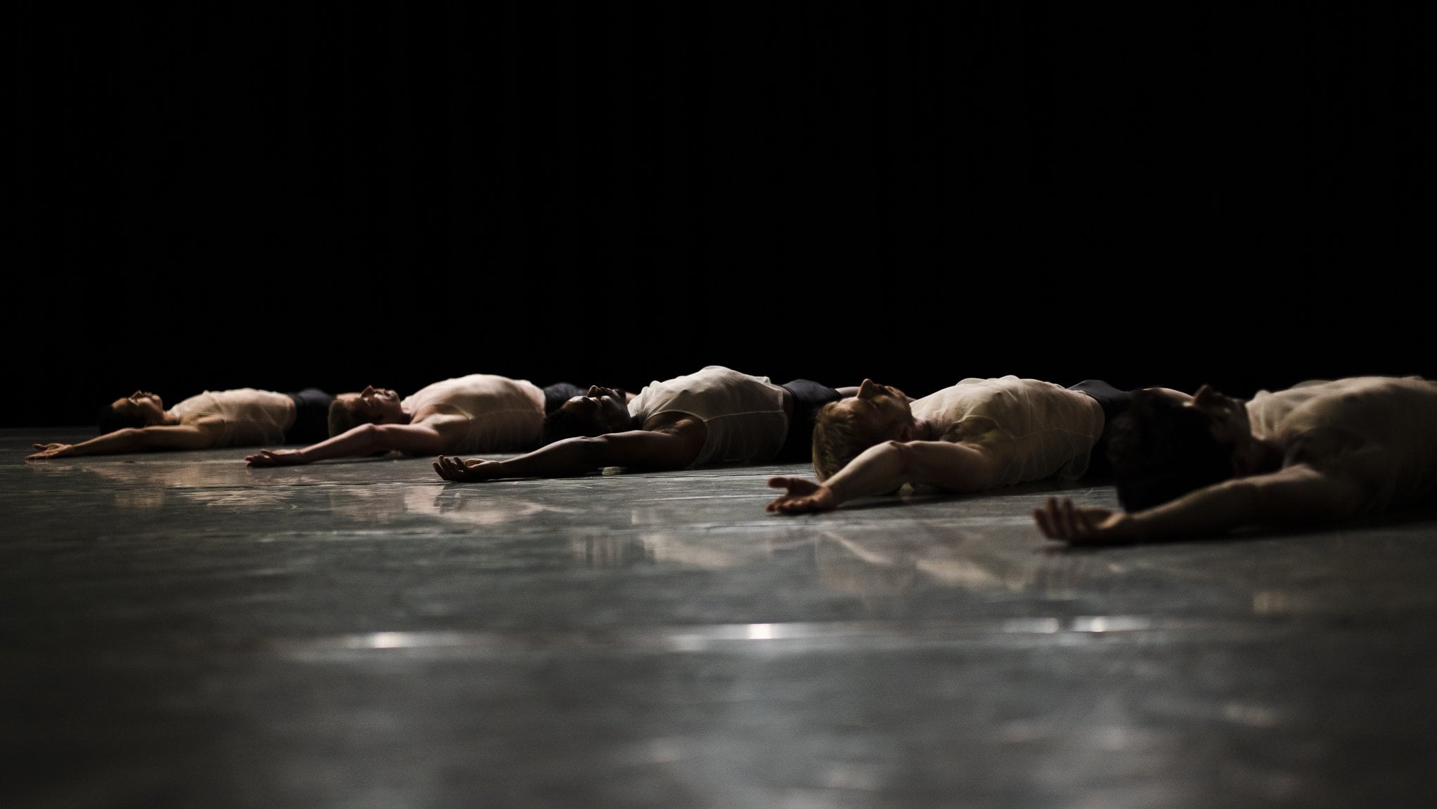 Dancers from the L.A. Dance Project lying on a black stage with their right arm raised.