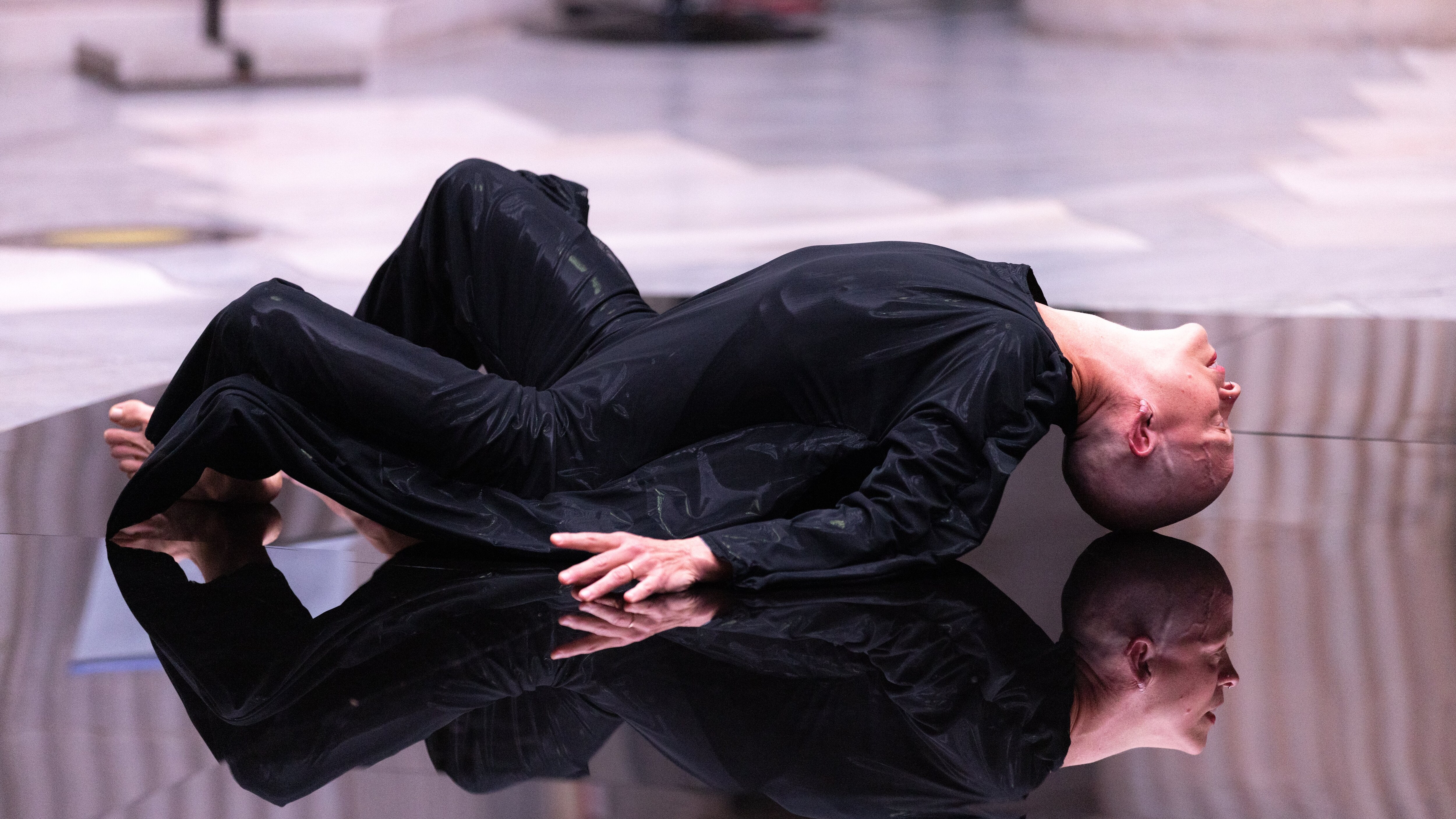 Bald woman dressed in black, lying on a reflective floor