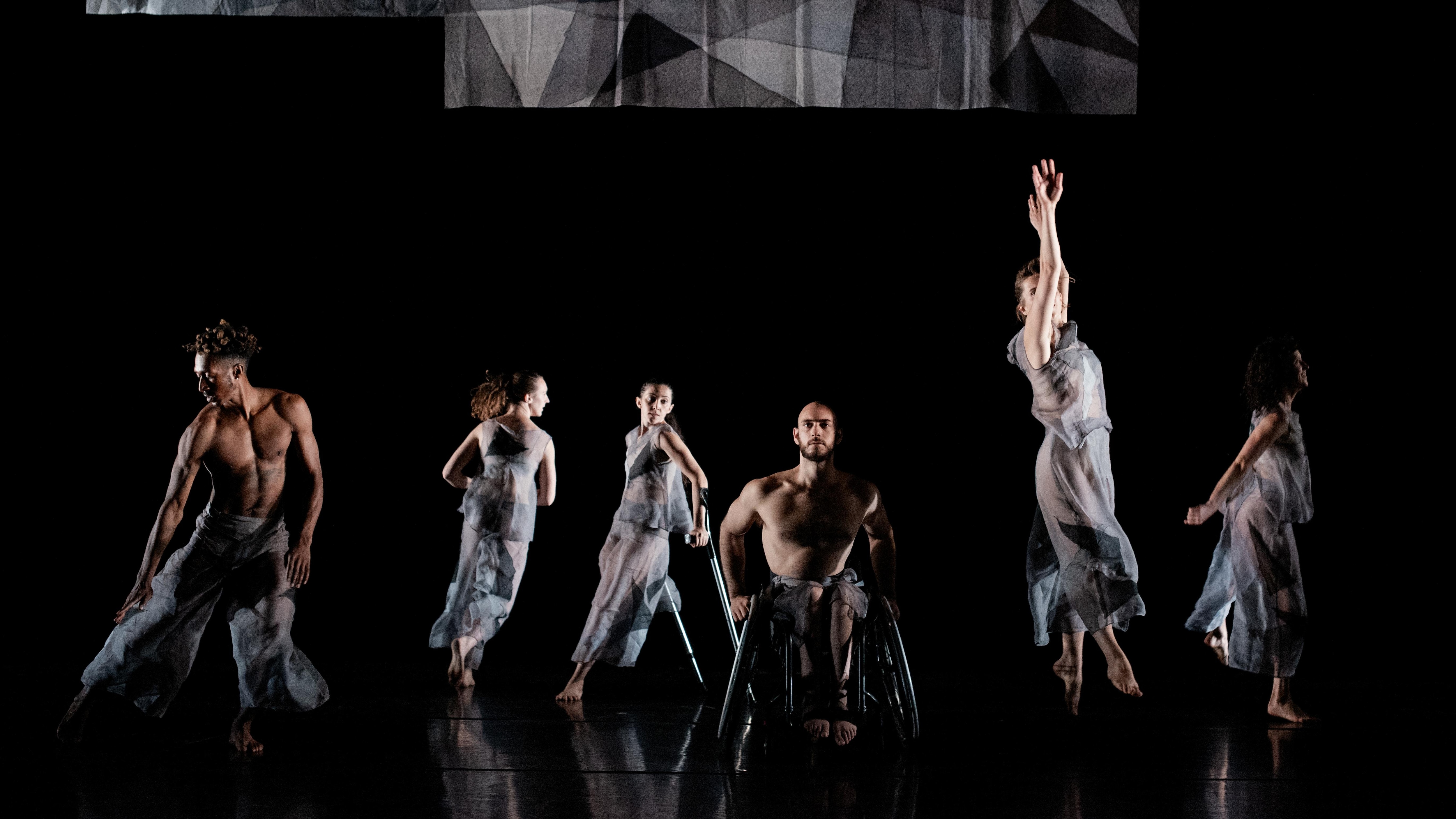 Six Dancers dressed in  see-through grey clothes dancing on a black stage
