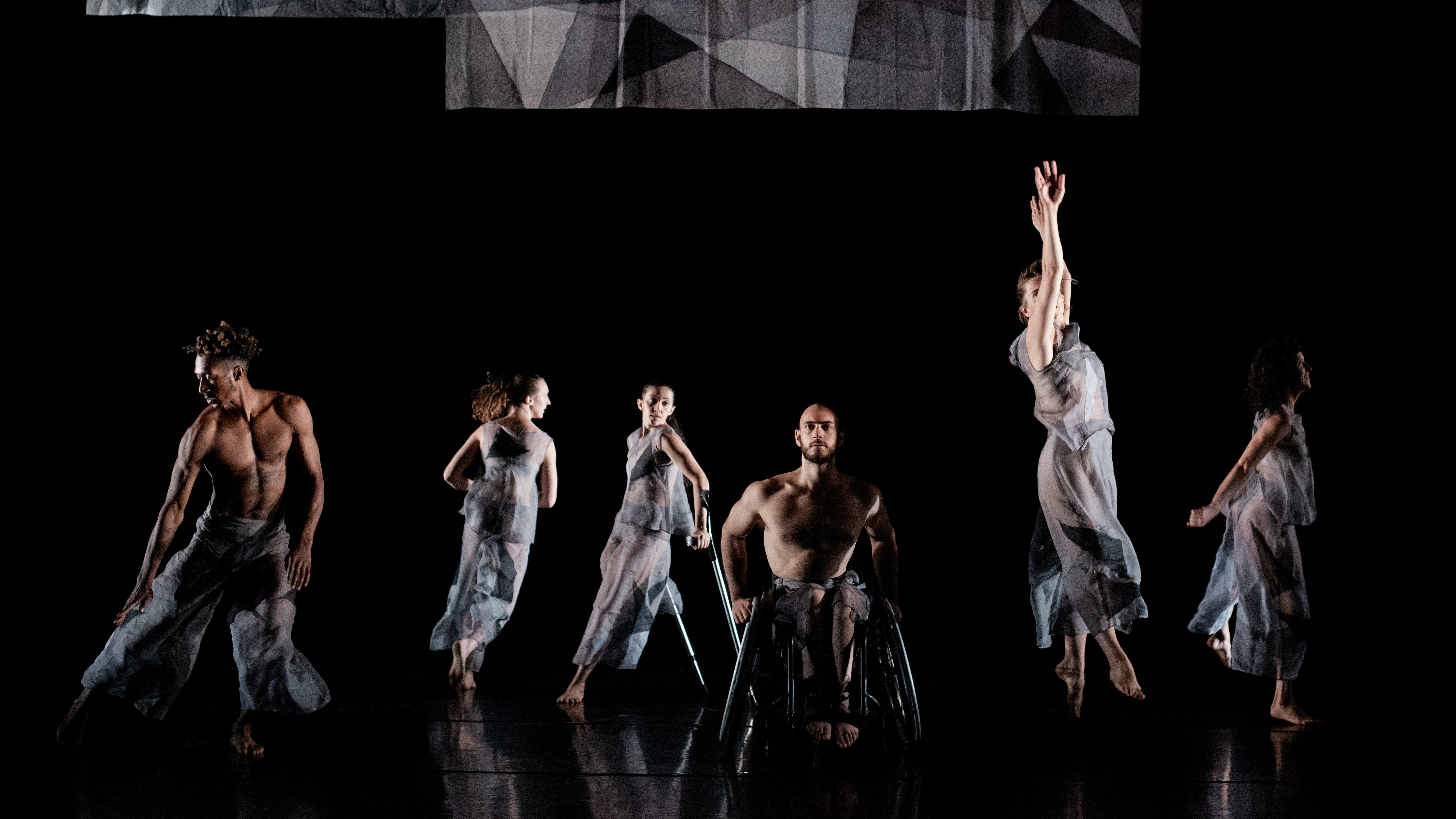 Six Dancers dressed in  see-through grey clothes dancing on a black stage