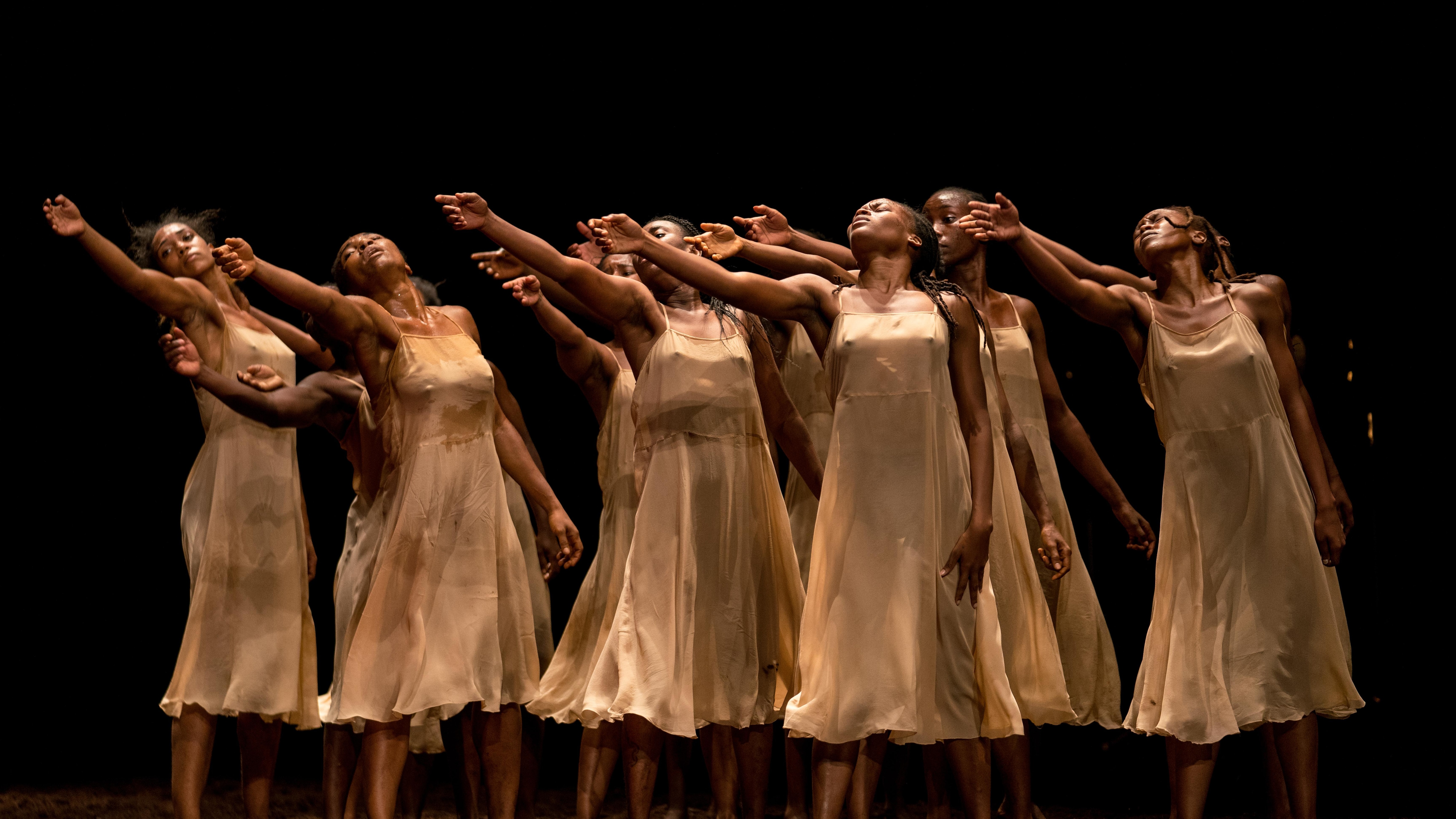 Women in white dresses, standing in dirt, facing three-quarters forward with their right arms raised.