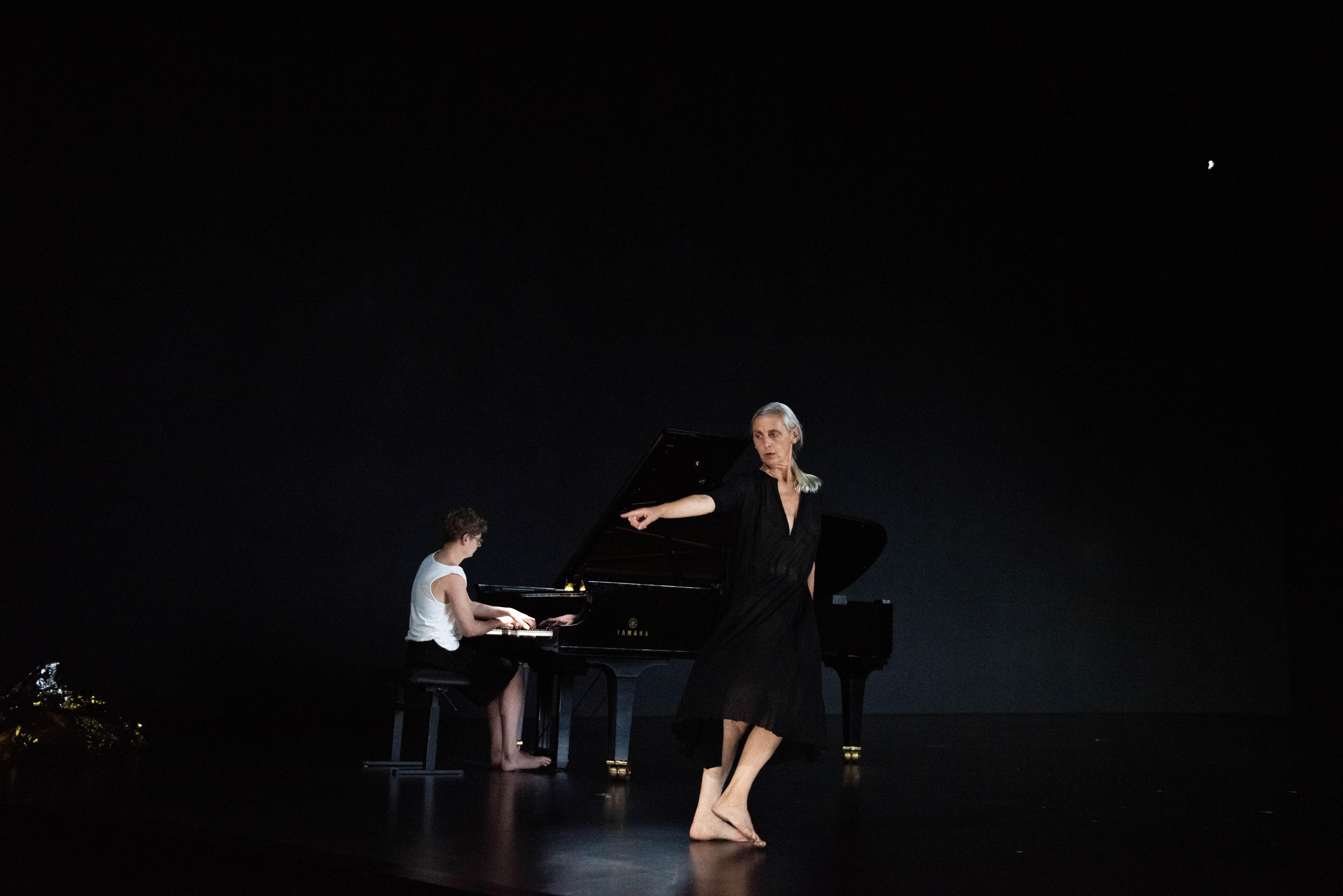 「在黑色的舞台上，身穿深V領黑色連身裙的舞者指向左方，她的赤腳引領著她朝相反的方向前進，而身後則是一名正在彈琴的青年。」