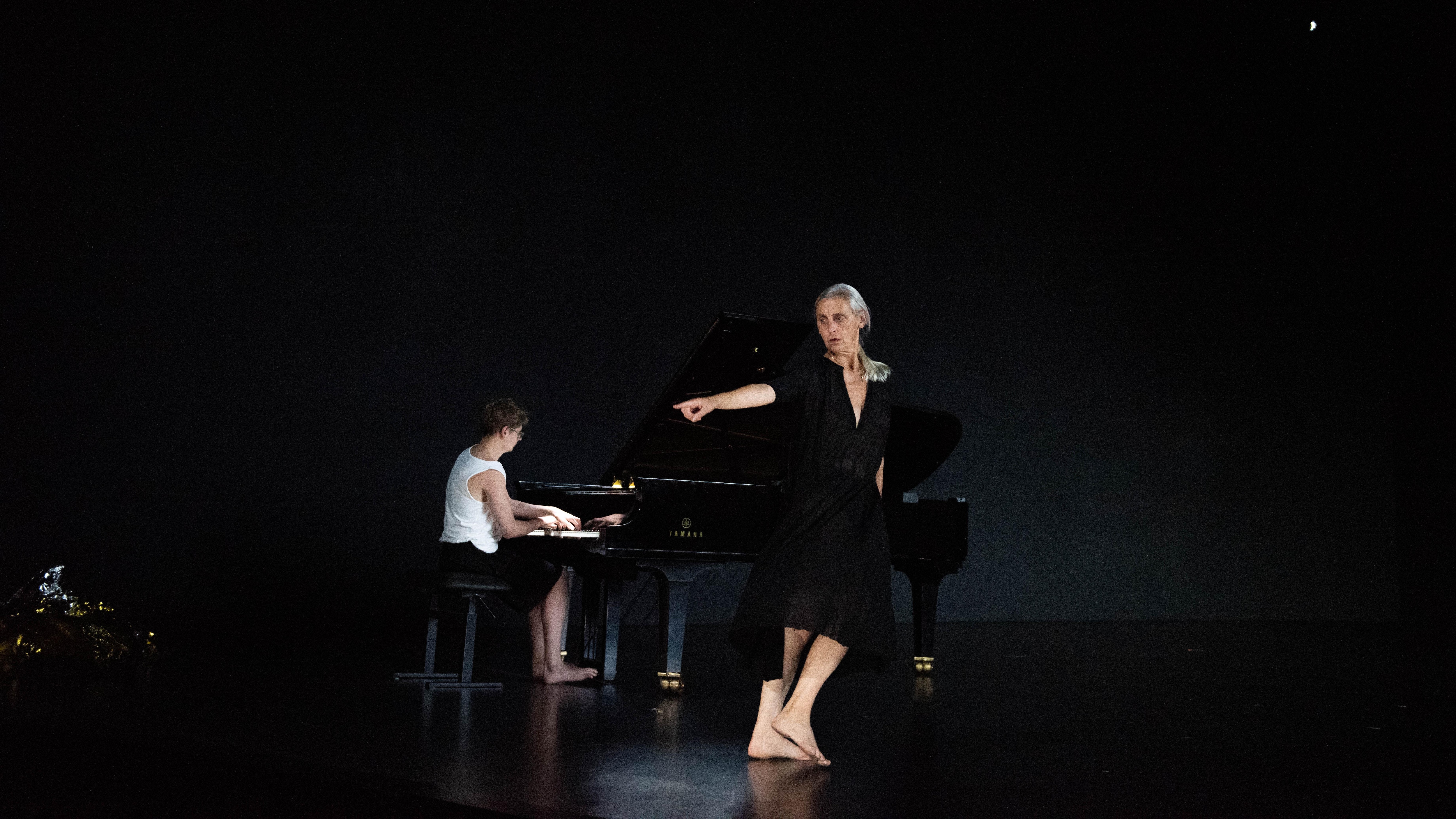 Sur une scène noire, une danseuse vêtue d’une robe noire avec un profond col en V pointe du doigt vers la gauche, ses pieds nus la menant dans la direction opposée avec, derrière elle, un jeune homme jouant du piano.