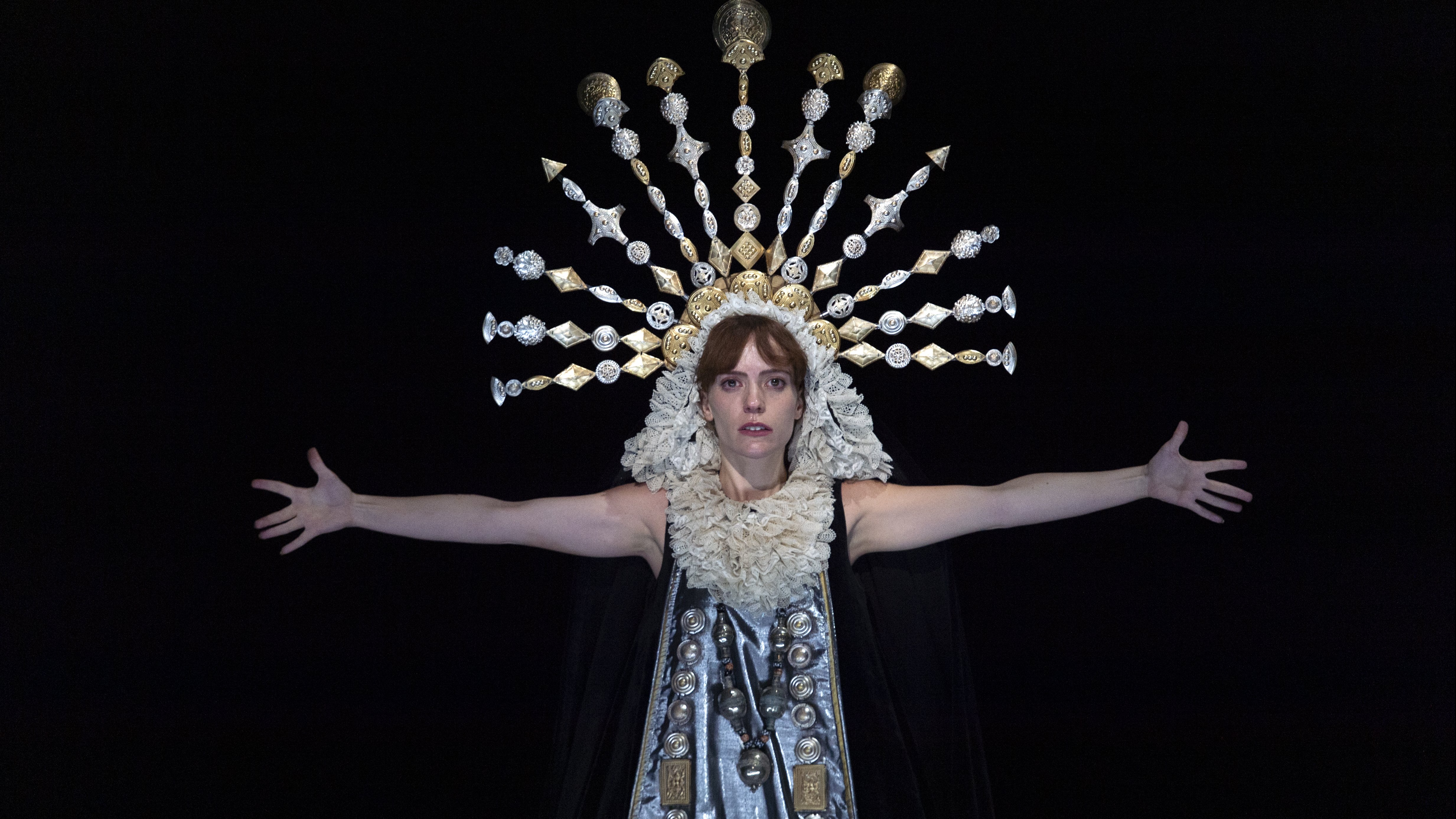 Woman in darkness whose face is bathed in light, wearing a crown in the shape of a halo, with her arms opened.  