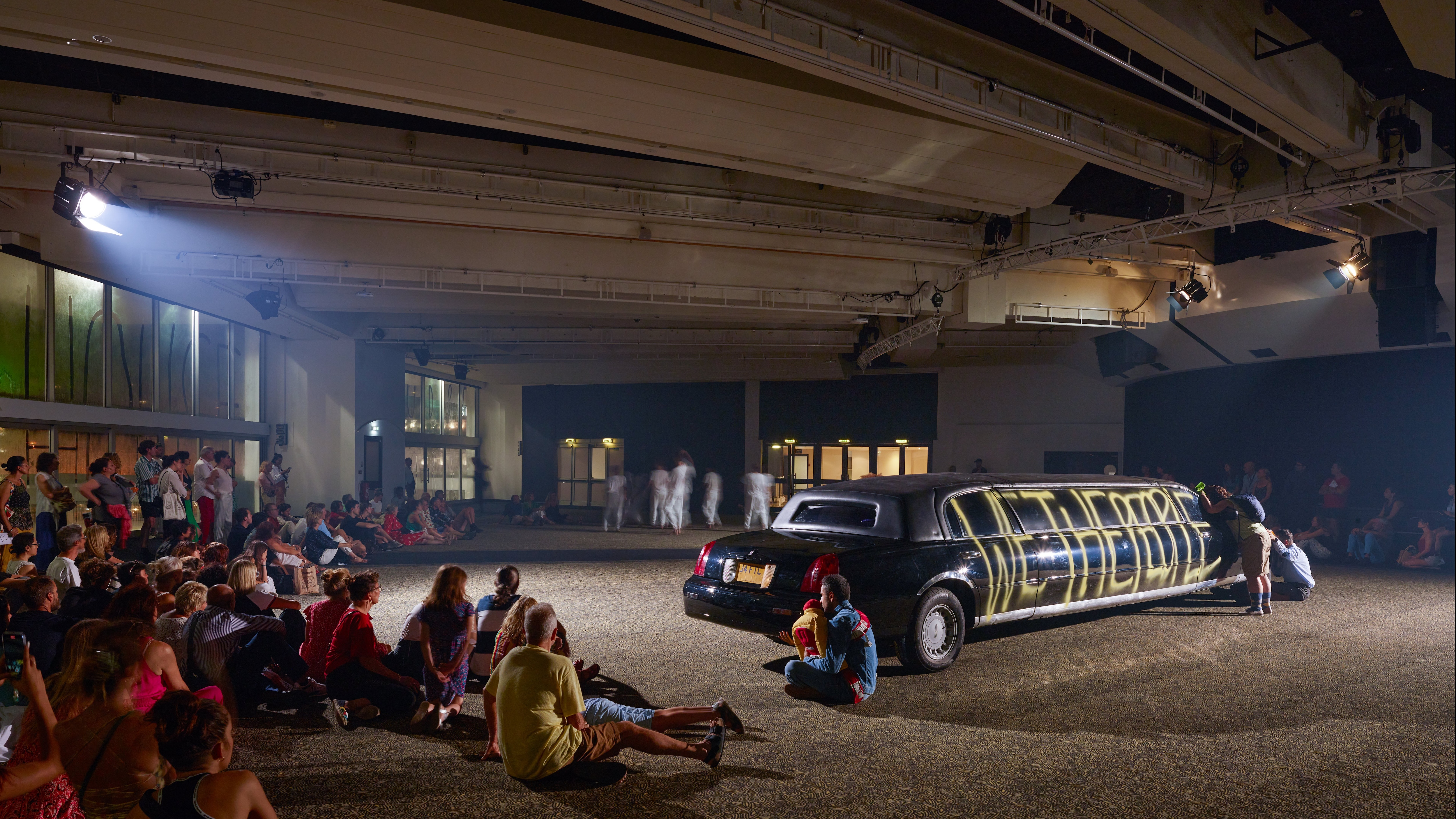 audience sitting on the floor in front of a black limousine