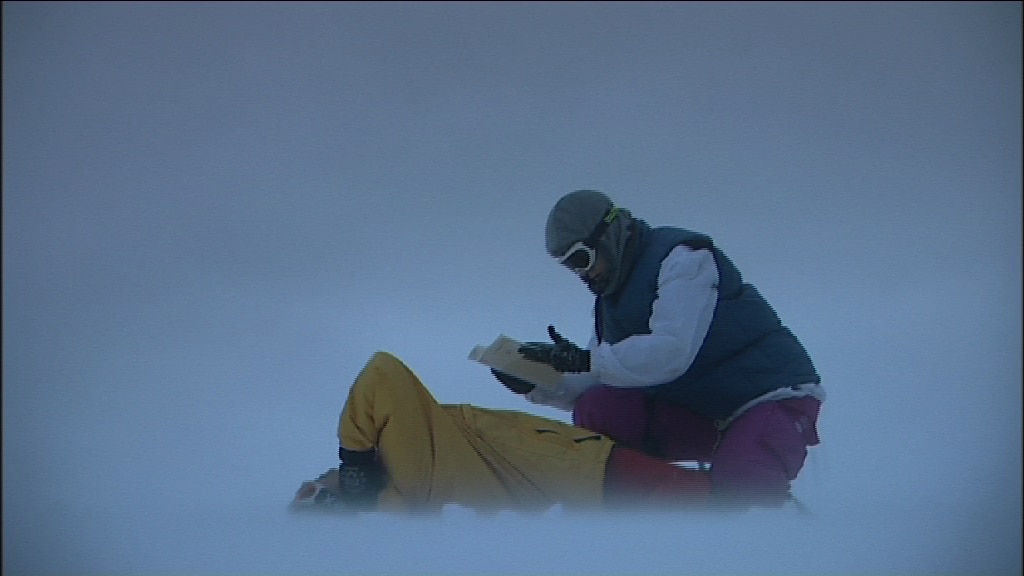 Two dancers on the floor, one kneeling over the other, wearing a mask and hood and holding a book.