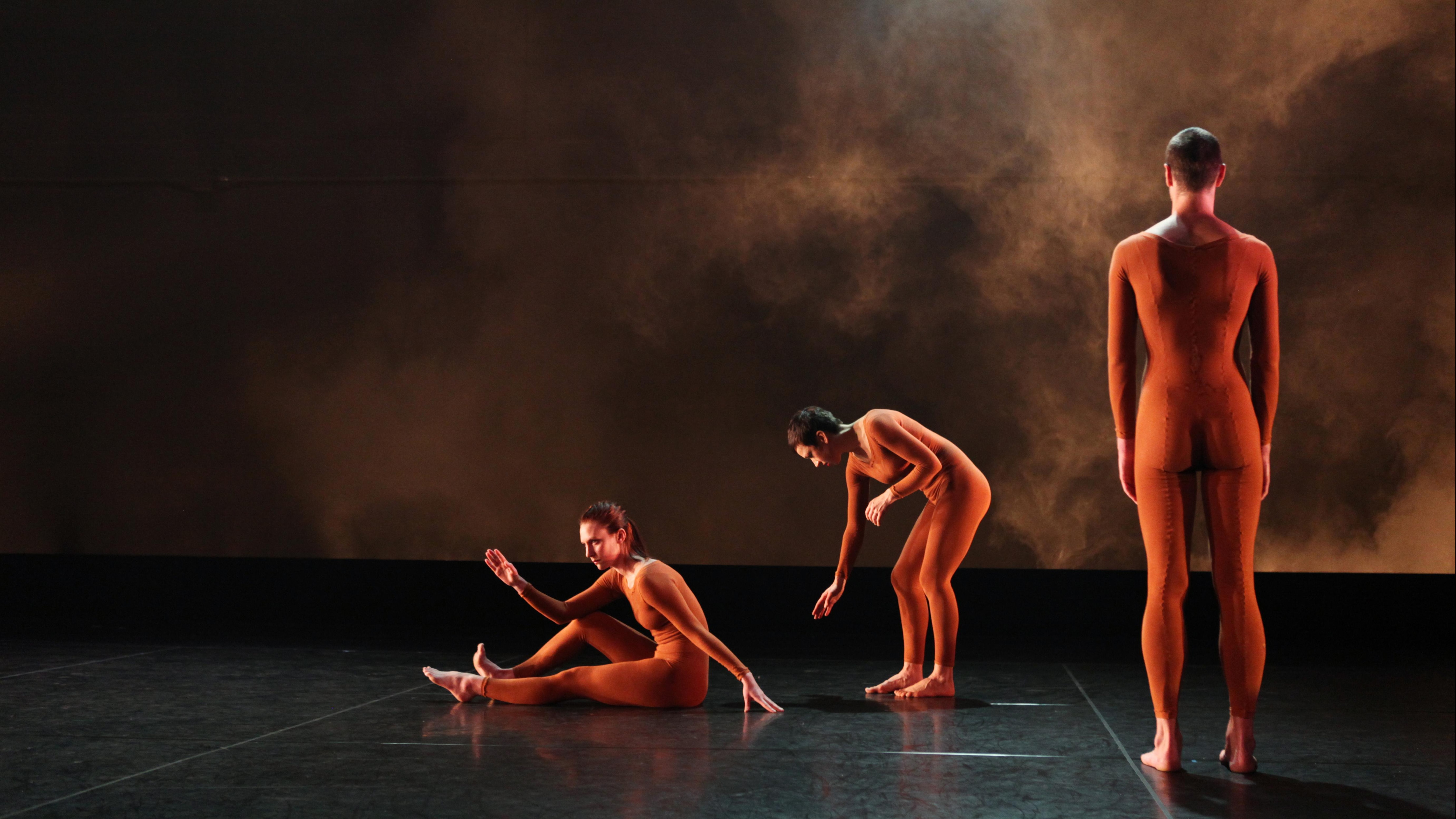 Three dancers on stage, the first one is sitting with his legs on the floor and one arm on his knee, the second one is leaning down, and the third one is standing from behind