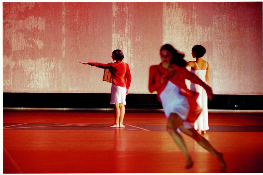 Trois danseuses sur scène, celle en premier plan court, les deux autres sont de dos en fond de scène, le bras gauche tendu à l’horizontal