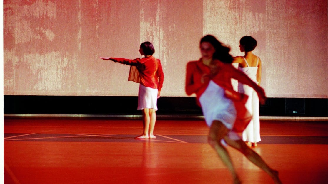 Trois danseuses sur scène, celle en premier plan court, les deux autres sont de dos en fond de scène, le bras gauche tendu à l’horizontal