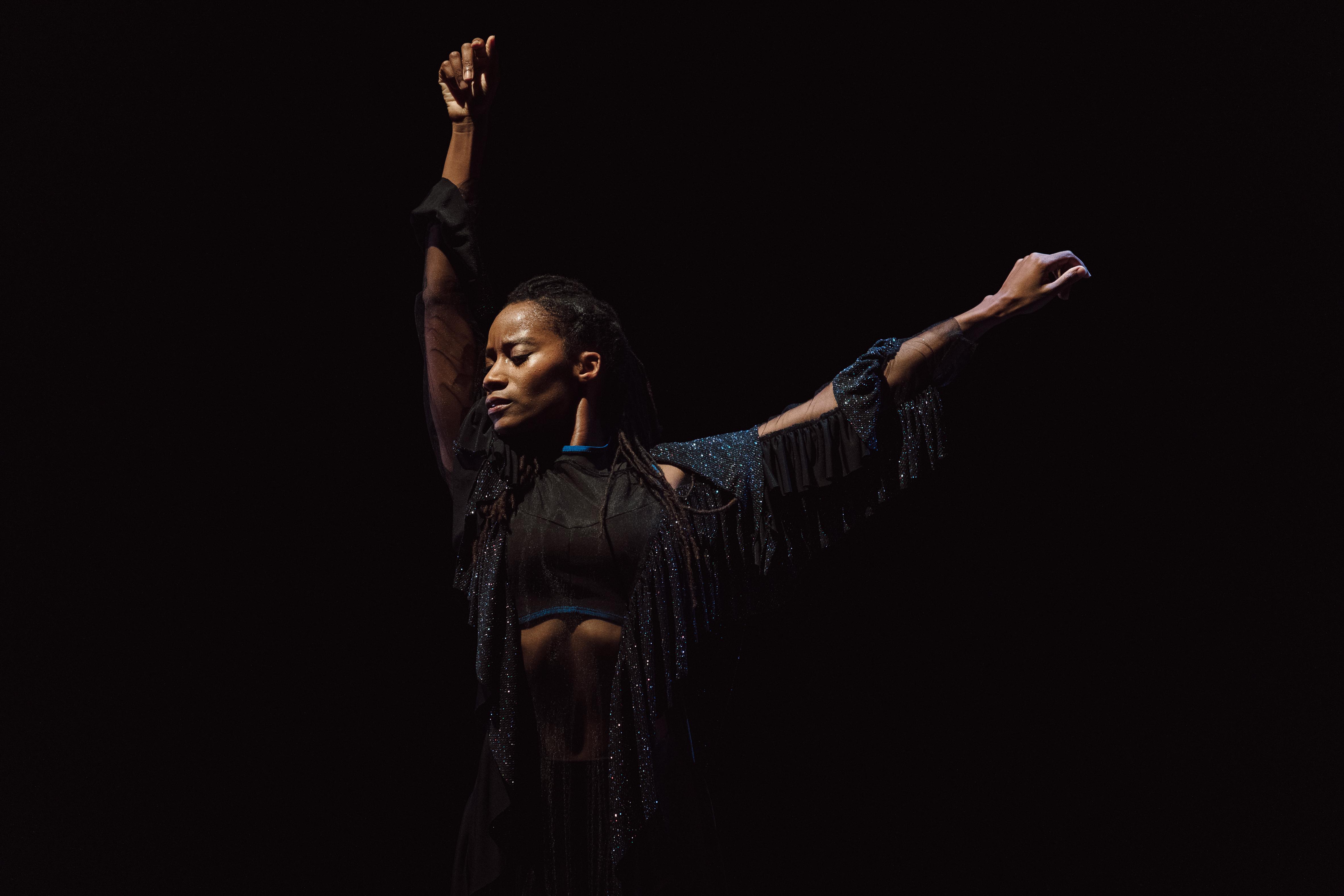 Danseuse vêtue de noir avec les bras tendus vers le ciel dans Mailles de Dorothée Munyaneza