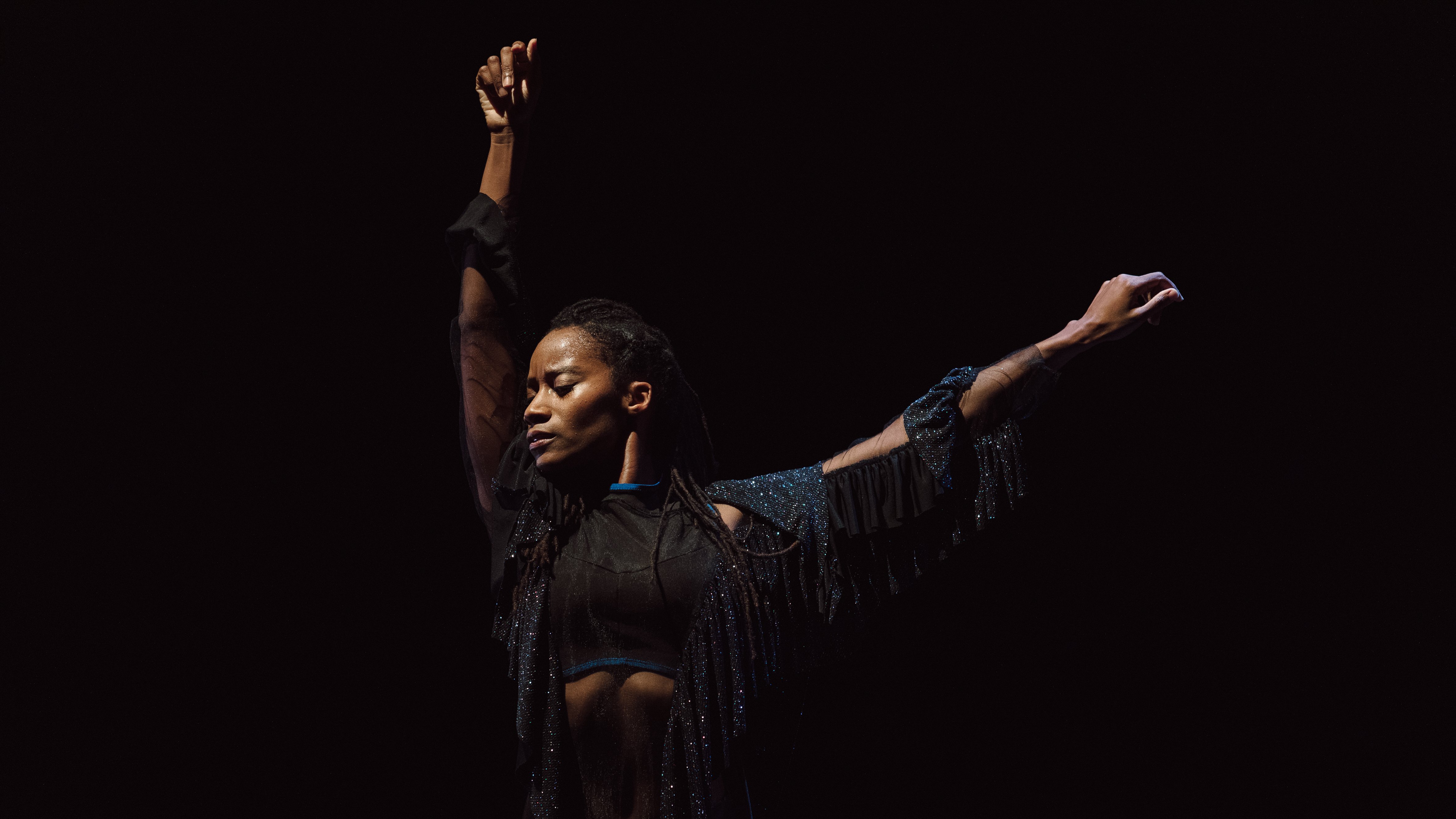 Danseuse vêtue de noir avec les bras tendus vers le ciel dans Mailles de Dorothée Munyaneza