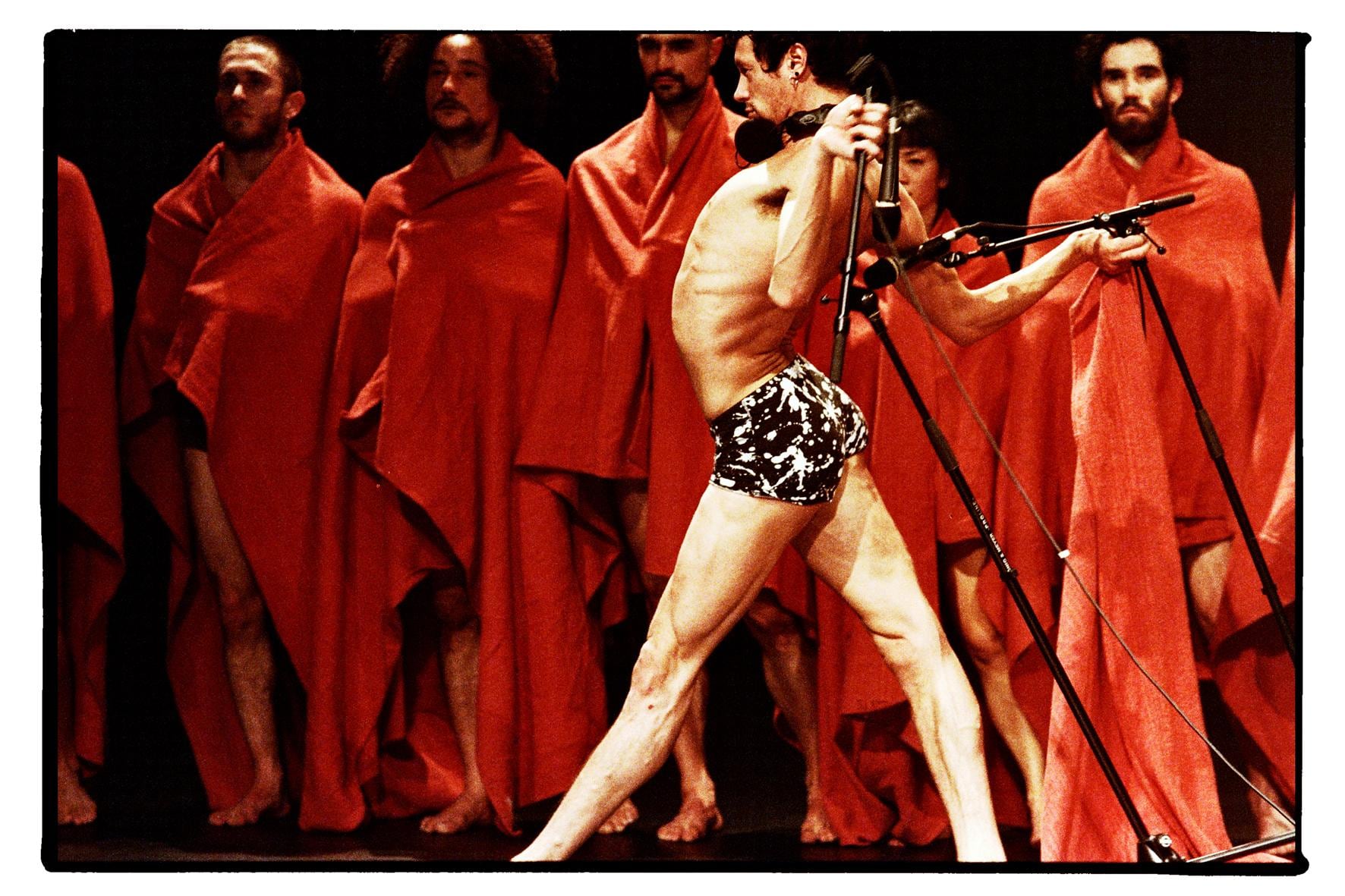A dancer in underwear, back arched, holding microphones behind him. He stands in front of people draped in red.