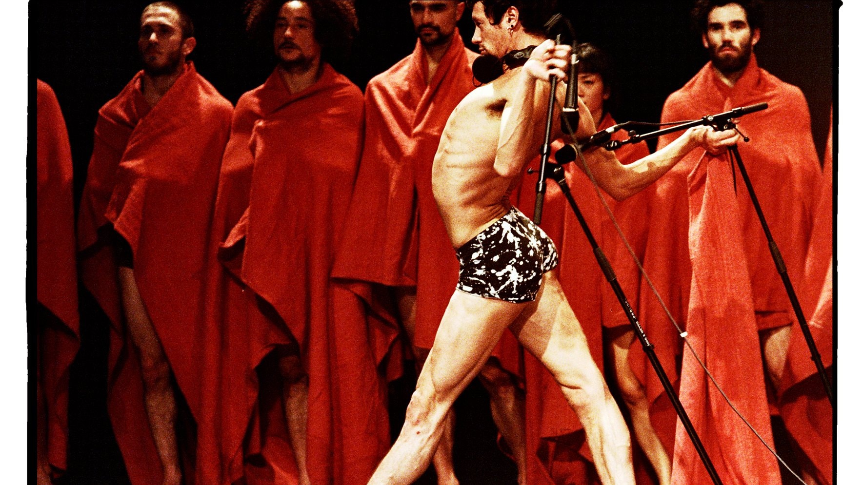 A dancer in underwear, back arched, holding microphones behind him. He stands in front of people draped in red.