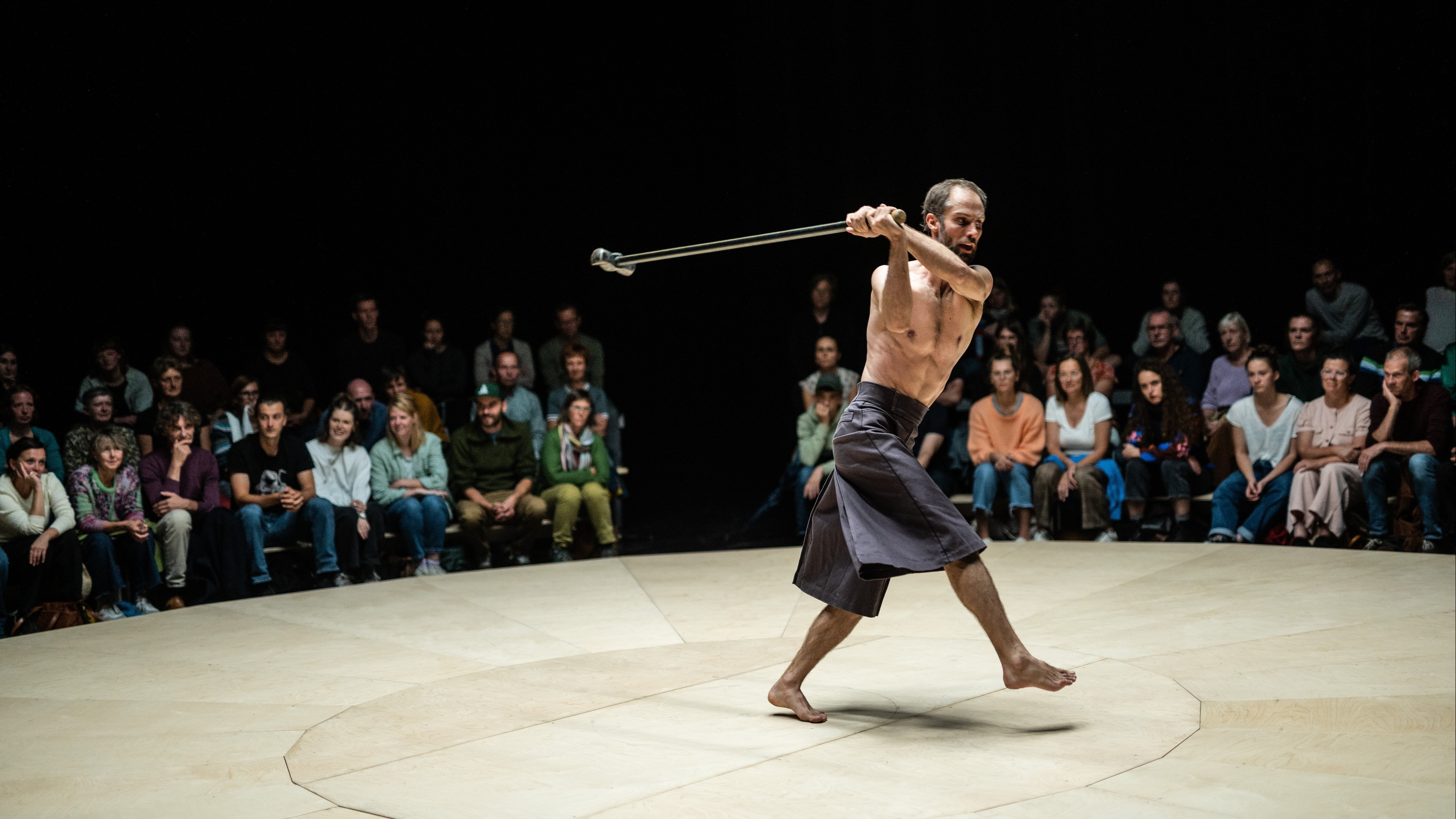 Bare-chested Alexander Vantournhout holding a big hammer and surrounded by the audience.