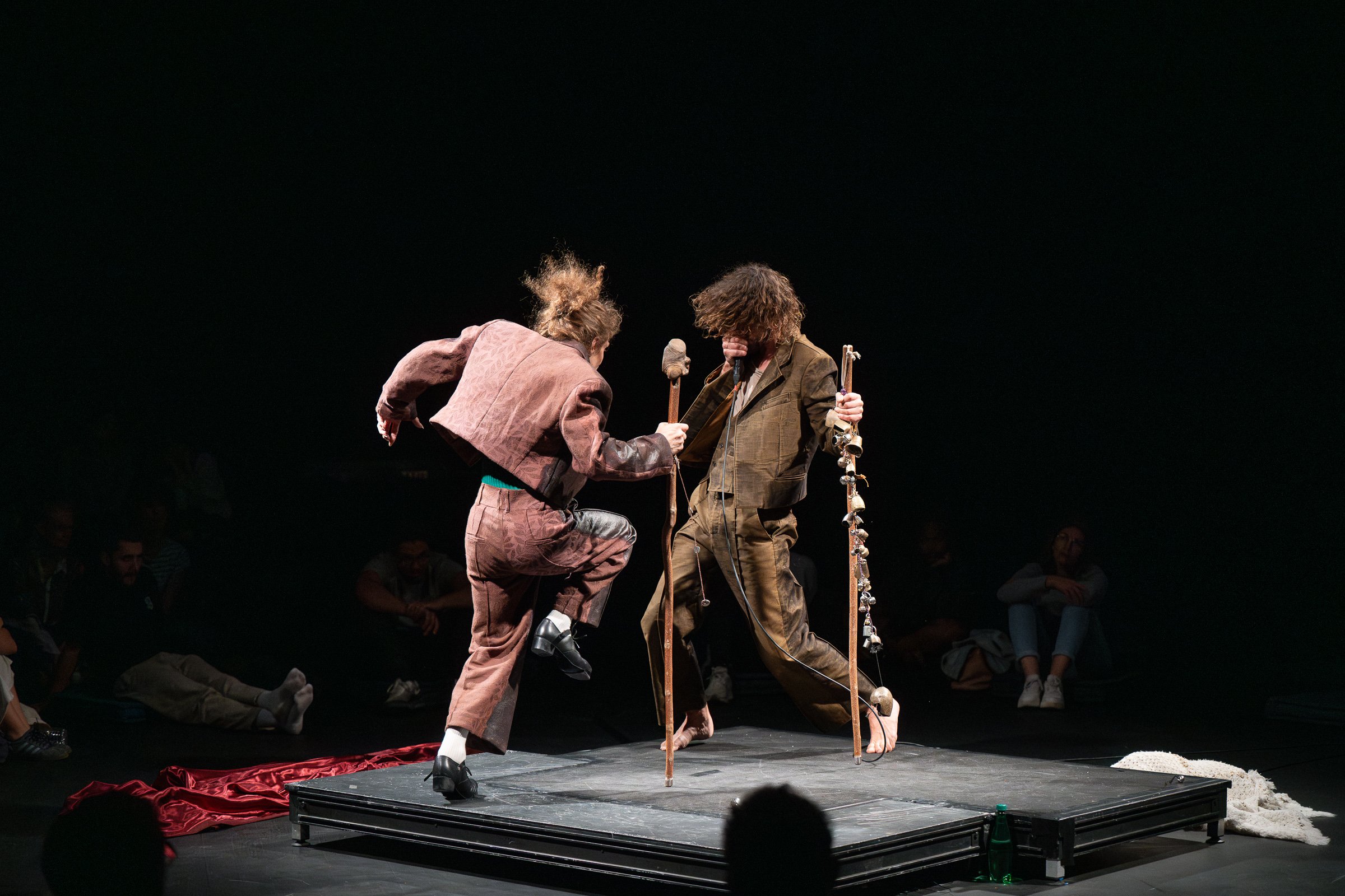 Two men in motion on a tiny square stage, holding sticks with bells.