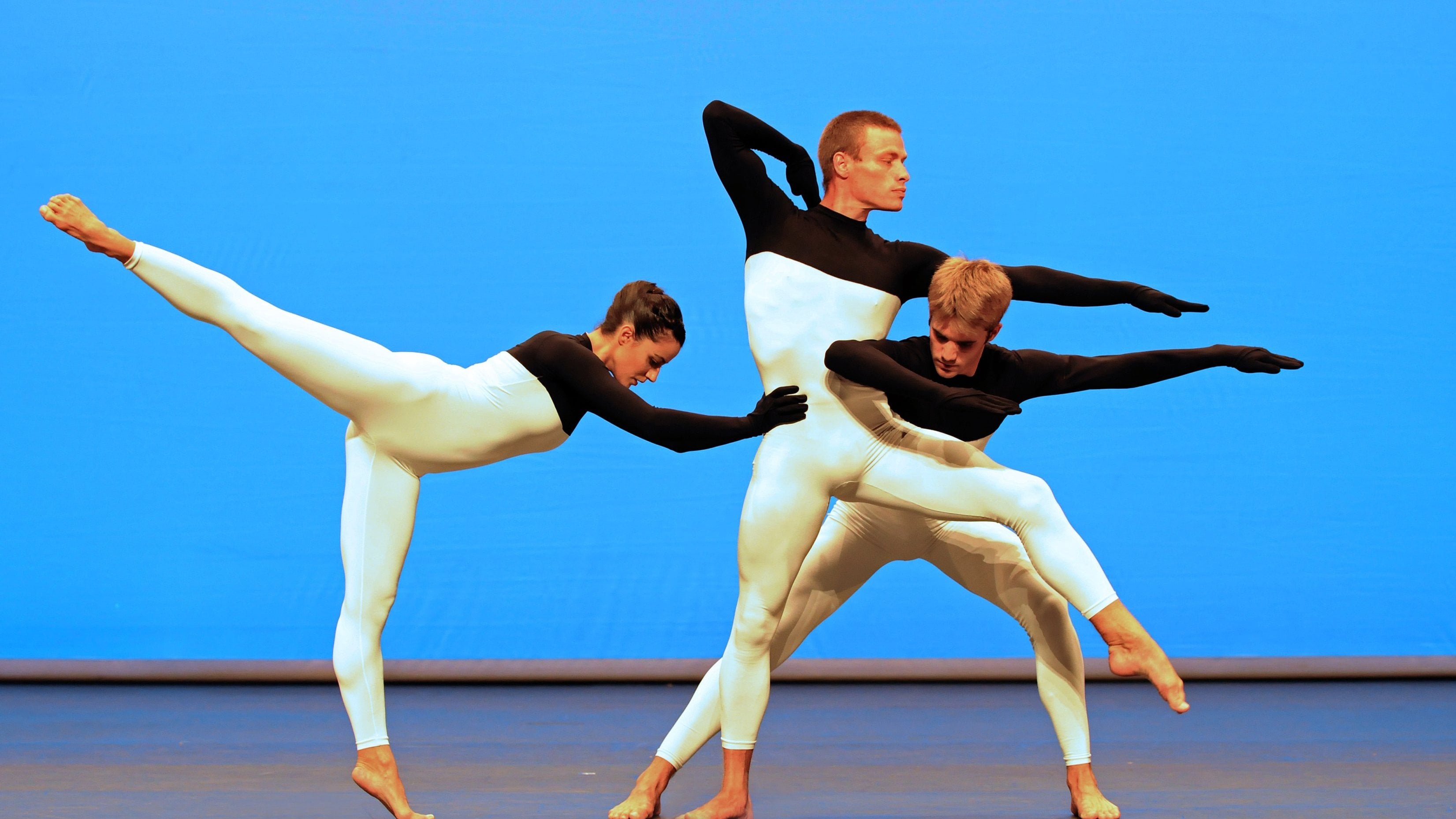 Trois danseurs en justaucorps longs noirs et blancs, devant un fond bleu. Extrait de Beach Birds. 