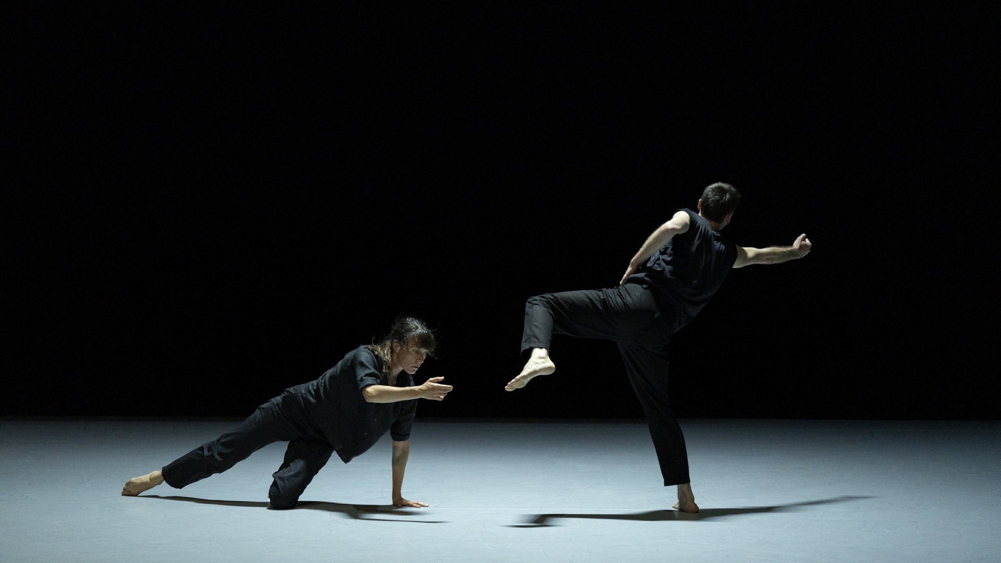  a woman on the ground and a man standing, dressed in black on a black background.