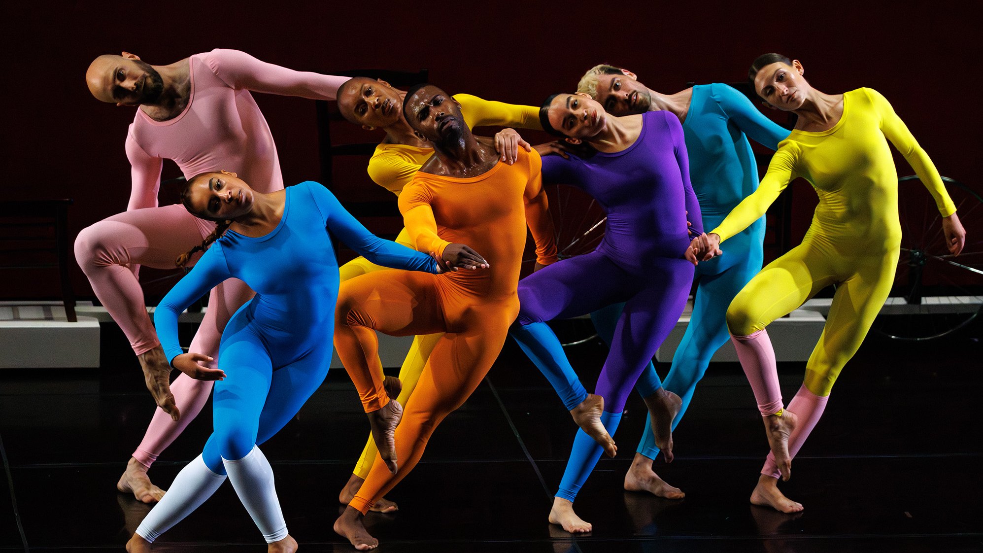Seven dancers dressed in single-colored, tight-fitting jumpsuits, all in the same position, standing on one leg with the other leg bent back.