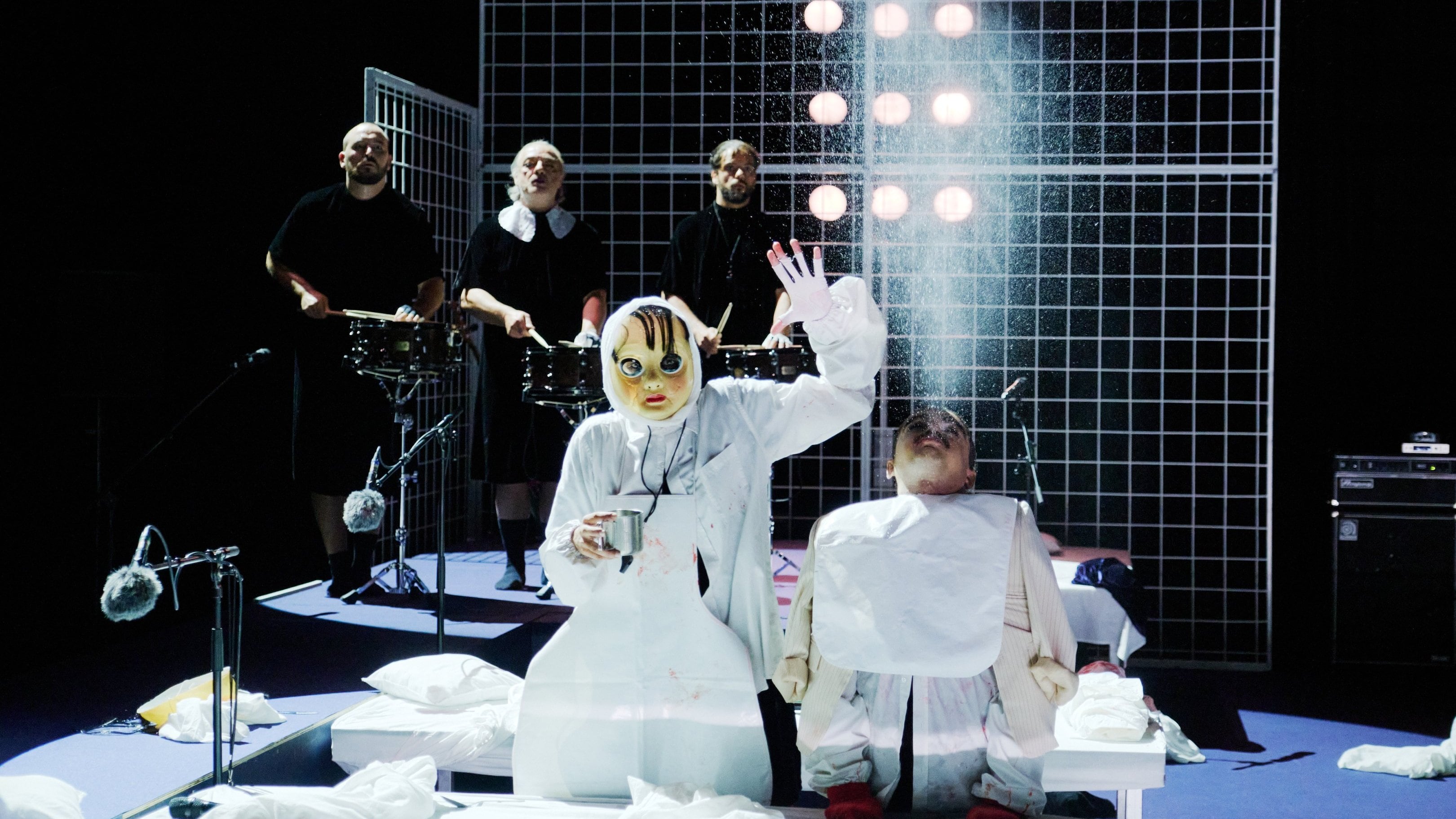 deux personnages en costumes blancs, l'un masqué et l'autre ressemblant à une marionnette, devant des musiciens