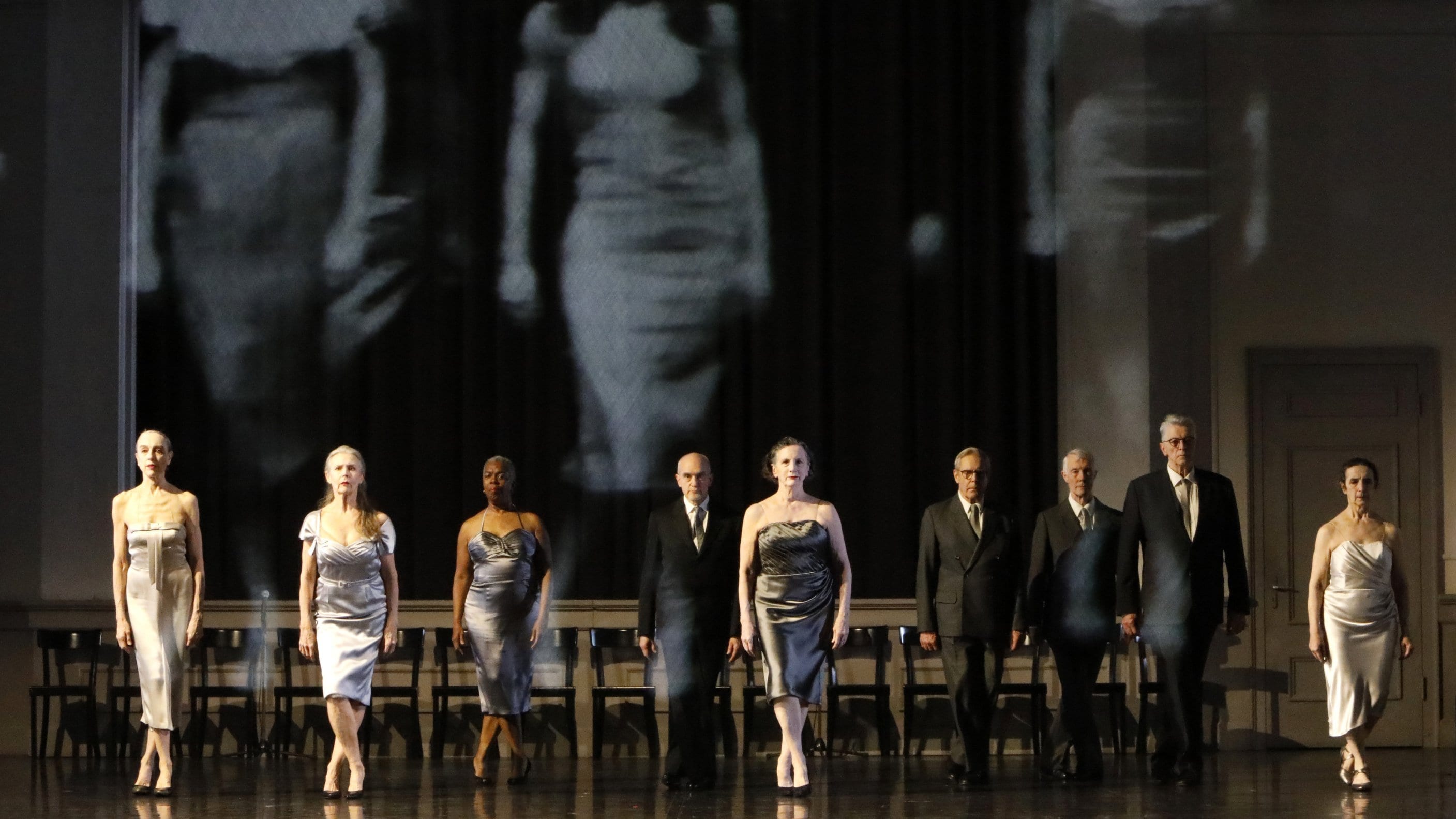 danseurs en tenue de soirée devant une projection en noir et blanc de l'ancienne version du spectacle