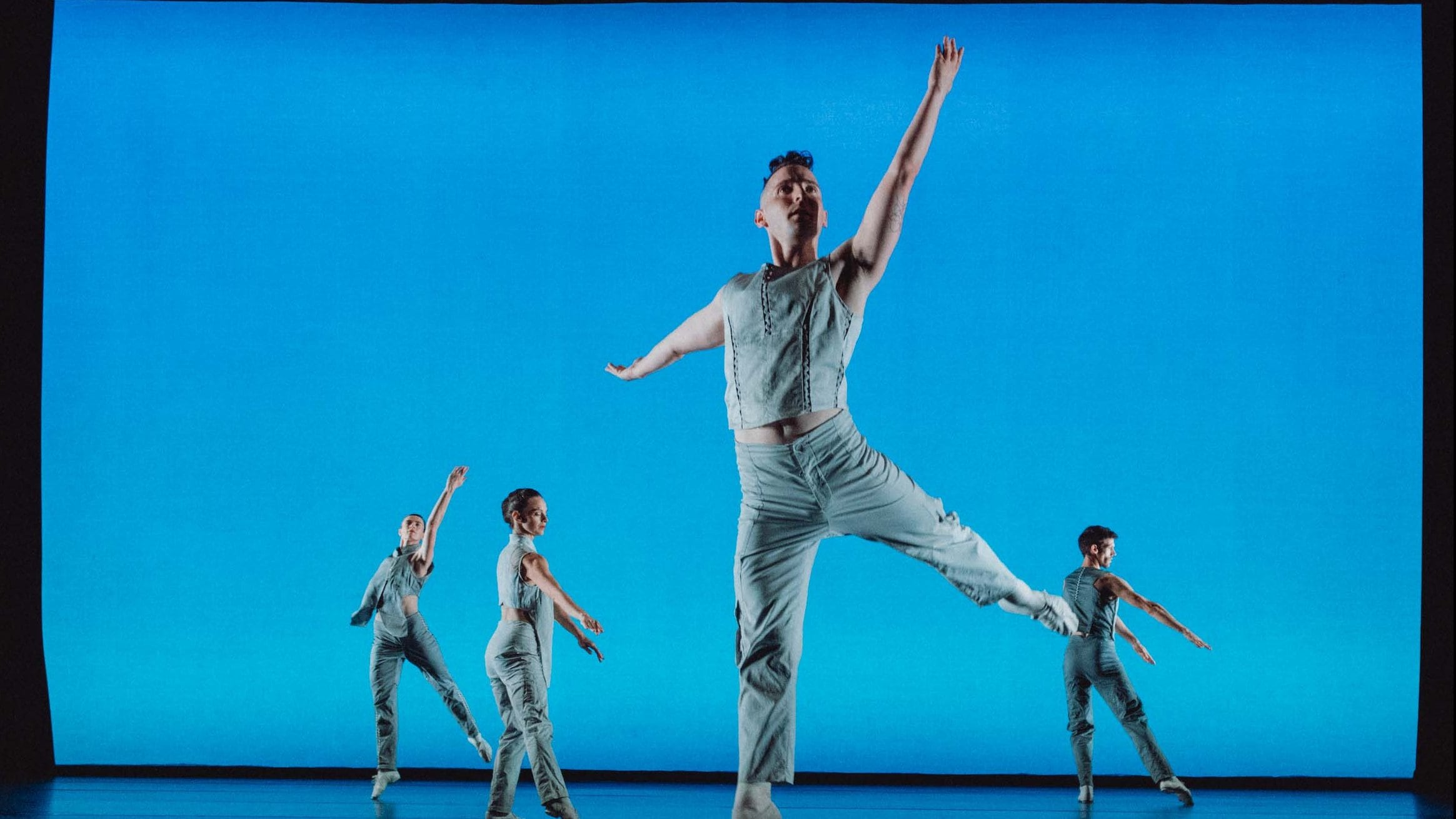 Four dancers in light-colored costumes in front of a vibrant blue background